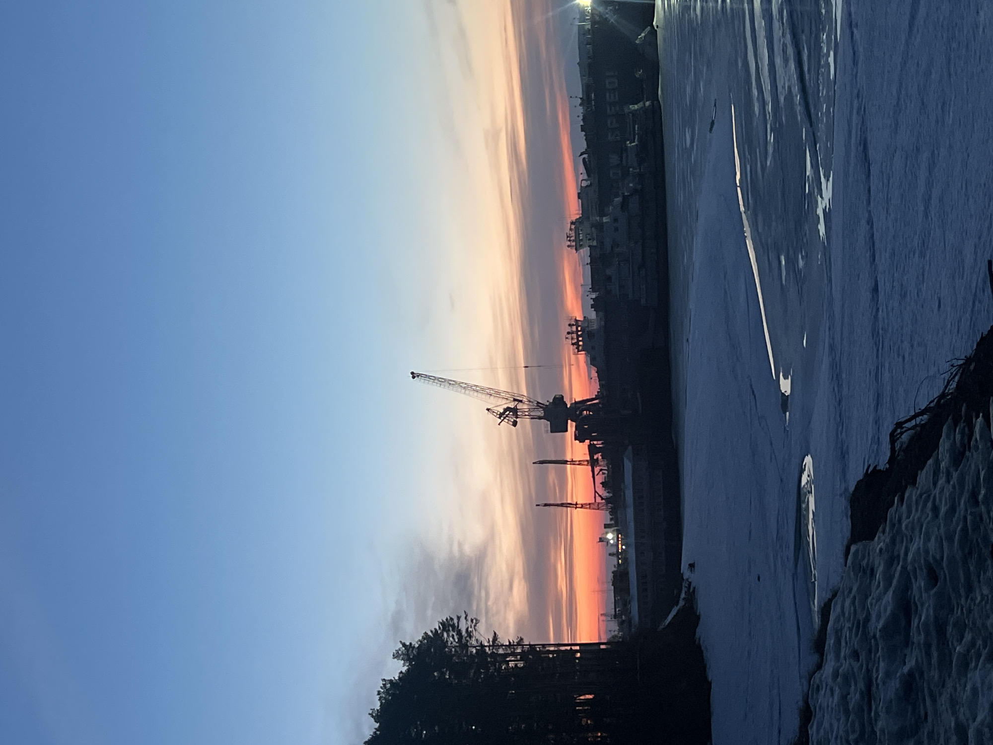 The image is a horizontally oriented, slightly low-angle shot of a twilight scene in what is likely the city of Chaikovsky, Russia. A sky gradient transitions from blue in the upper left to peach/orange/red on the right horizon. The prominent silhouettes of at least two cranes dominate the mid-ground, indicating a port or industrial area. The background reveals the faint outlines of buildings and lights, suggesting a cityscape. A dark line of trees is at the lower left, and a snow-covered bank or shore is in the lower right foreground. Patches of ice or water reflect light.