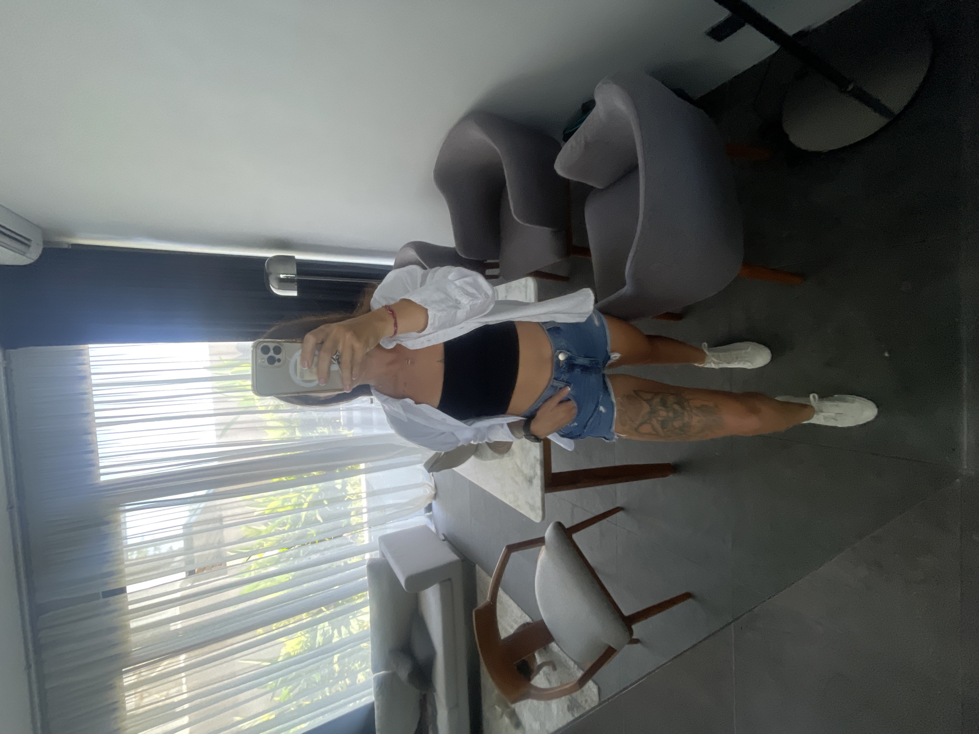 A woman is standing in a room, taking a mirror selfie. She is wearing a black crop top, a white open shirt, denim shorts, and white sneakers. She has a tattoo on her left thigh. The room appears to be a living area with a sofa, chairs, and a dining table. Large windows with sheer curtains are visible, letting in natural light. The floor is dark tile. A floor lamp is also present in the room. The context of Kediri, Indonesia is not discernible from the visual information provided.
