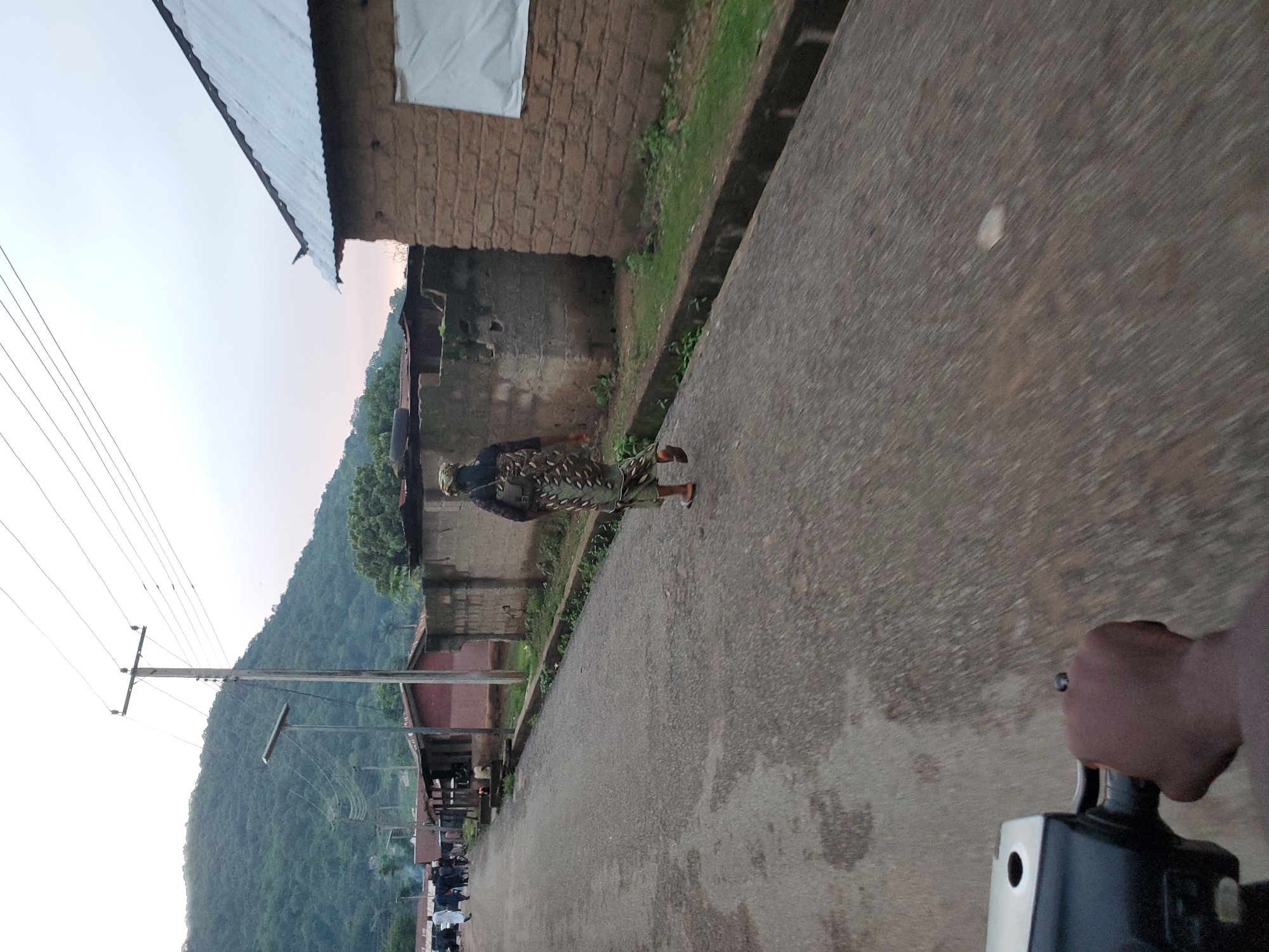 The image captures a street scene in Mararaba, Nigeria, from a low-angle perspective, likely from a moving motorcycle, indicated by a dark-skinned hand gripping a black and silver handlebar in the lower-left foreground and motion blur on the road surface.
A lone woman, dressed in a patterned green, brown, and white long garment and sandals, walks away from the viewer in the center of the asphalt road, carrying a light-colored backpack. The road itself is gray, with visible aggregate and some lighter patches, extending into the distance.
Along the right side of the street, a narrow concrete curb separates the road from an unpaved strip of sparse green grass and dirt. Beyond this strip are multiple single-story structures with rough-textured, earth-toned mud-brick walls and corrugated metal roofs. One building on the far right features a white, covered window opening. Adjacent to it, a segment of a concrete block wall is visible.
On the left side of the street, a similar setup of a curb, green vegetation, and structures is present. A tall wooden utility pole supporting multiple overhead power lines stands prominently. Next to it, a modern-looking street light pole with a solar panel on top is visible. Further down the street on the left, a cluster of individuals dressed in various garments and several parked vehicles can be discerned.
The background is dominated by a large, green, tree-covered mountain range or hill. The sky above is overcast and pale, transitioning to a very faint pinkish-orange hue closer to the distant horizon.