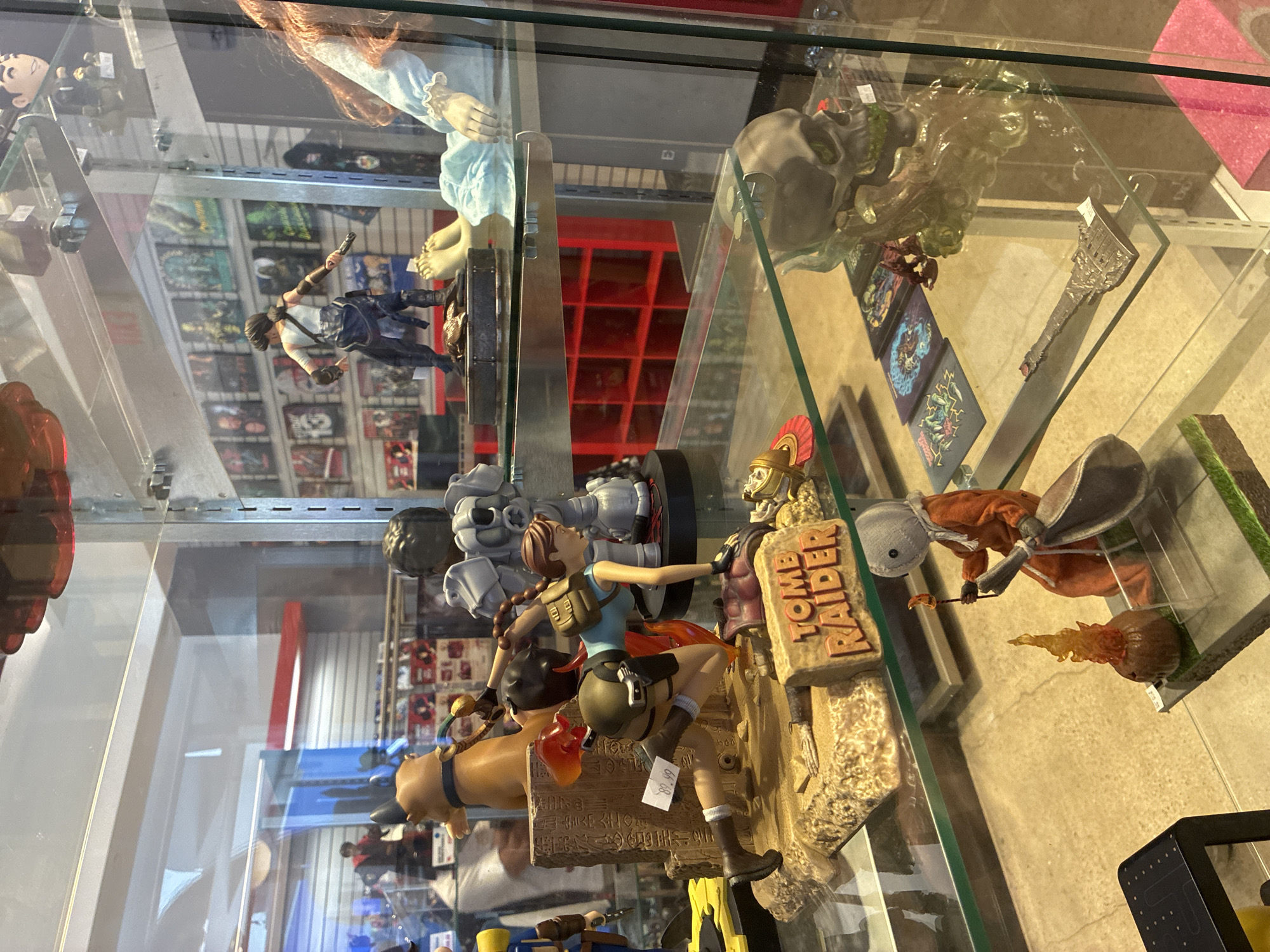 In an indoor display case, a variety of collectible figurines and merchandise are showcased. The primary focus is on a "Tomb Raider" themed display featuring a Lara Croft figurine in an action pose, an artifact, and the "Tomb Raider" logo. Adjacent to this, a stylized skull figurine with moss-like accents is positioned on a shelf. To the left, a figure resembling Sam from the movie "Trick 'r Treat" is depicted with a campfire accessory. Further back, other figures and media, possibly video games or movies, are visible on shelves, creating a densely populated retail display.