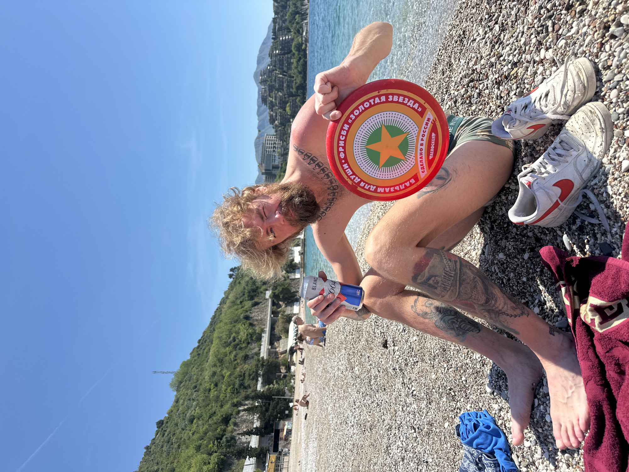 A man with a blond beard and curly hair, covered in tattoos on his arms, chest, and legs, is sitting on a pebbly beach in Budva, Montenegro, under a clear blue sky. He is holding a red frisbee in his right hand and a blue and red drink can (Blanc 1664, 500ml) in his left hand. He is wearing green camouflage-patterned shorts that say "VE INTIM". His white Nike sneakers are visible next to a red towel and a blue garment on the pebbles.
The frisbee is red with a circular design featuring a green and orange star. Visible text on the frisbee includes "ФРИСБИ «ЗОЛОТАЯ ЗВЕЗДА»" (Frisbee "Golden Star"), "БАЛЬЗАМ ДЛЯ ДУШИ" (Balm for the soul), "СДЕЛАНО В РОССИИ" (Made in Russia), and "AEROCKER".
The scene depicts a sunny day with bright light. In the background, there is clear blue sea water meeting the pebbly shore. Beyond the water, a lush green, hilly coastline rises, dotted with numerous buildings and resorts, indicative of a popular coastal area. Other beachgoers are visible further down the shore, lounging on the pebbles. The man appears to be relaxed, enjoying the beach environment.