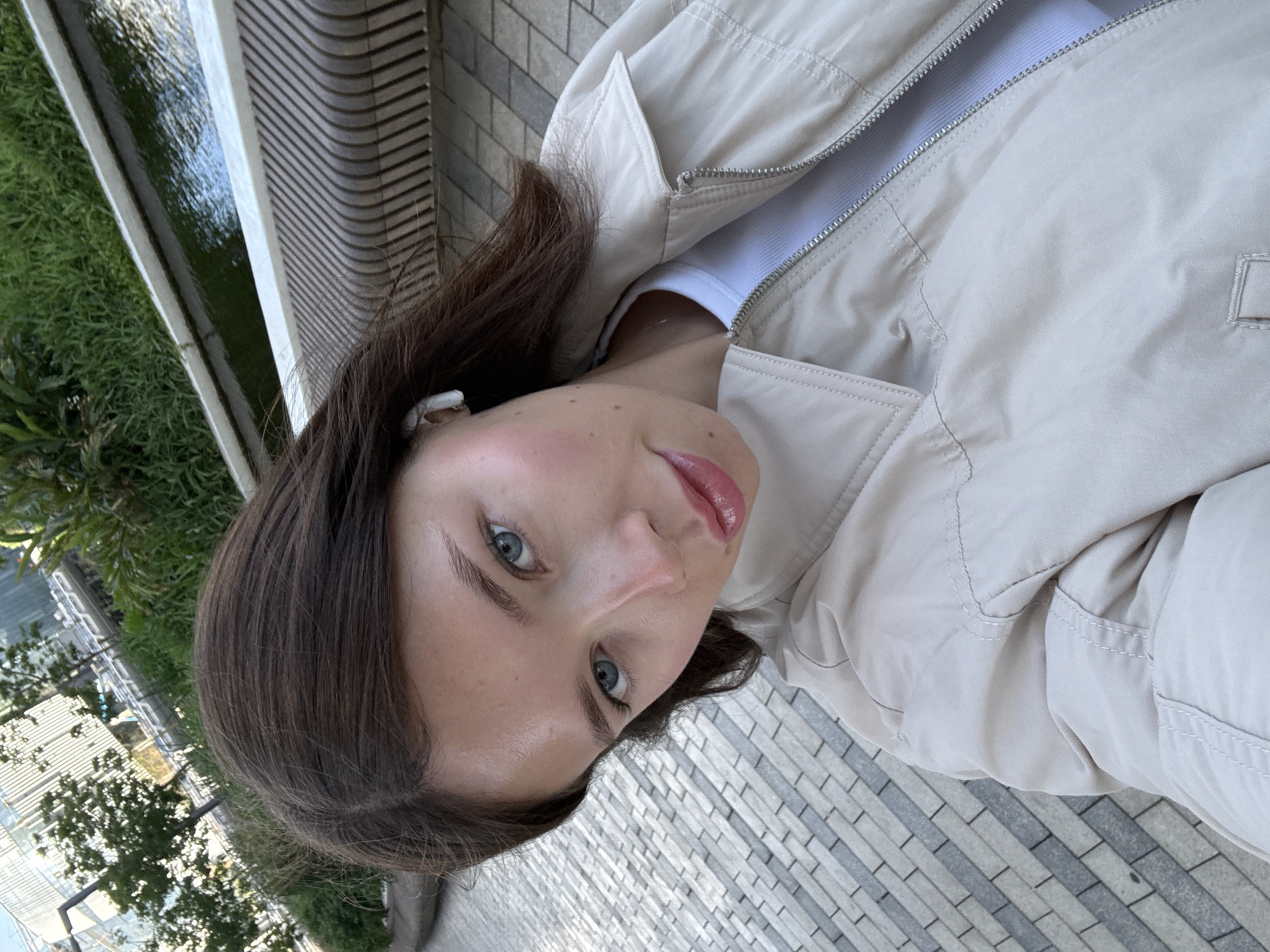 A woman is shown in a close-up, tilted selfie. She has brown hair, fair skin, and blue eyes. She is wearing a light-colored jacket and a white shirt underneath. An earbud is visible in her left ear.
The background includes greenery, water, and modern architecture. The patterned ground suggests a public space or walkway. Although identification is not possible without additional information, the location is stated to be Tel-Aviv, Israel.
