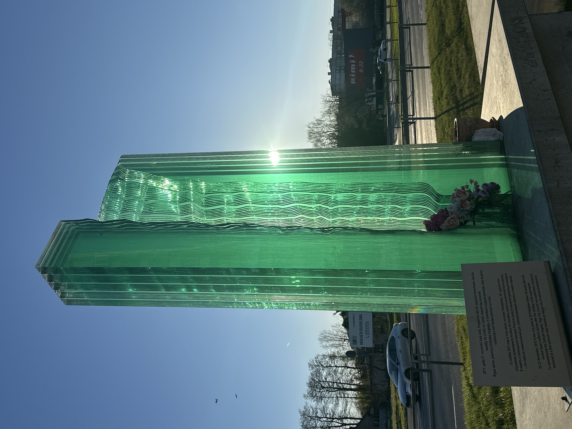 A striking green-tinted glass sculpture, composed of stacked and layered panes, stands prominently outdoors under a clear blue sky. The layers of glass exhibit a wavy texture, particularly in the upper sections, giving the appearance of flowing water or ice. The sun's rays cause bright reflections and flares on the glass, indicating bright daylight, likely early morning or late afternoon, as the sun appears low.
The sculpture serves as a memorial, with a white informational plaque placed at its base. Dried or artificial flowers in shades of pink, purple, and white are neatly arranged on the base next to the plaque, along with a terracotta pot and a piece of white fabric. The plaque, presented in Latvian, Russian, and English, details the tragic event it commemorates: "On 21 November, 2013 in this very place at 20 Priedaines Street, 54 people died and several dozen suffered injuries after the collapse of the supermarket. To commemorate the Zolitude tragedy, Riga City Council has established a temporary memorial as a first symbolic step towards an eternal site of national remembrance." The date "21.11.2013" is also carved into the concrete base.
In the background, a road is visible with some traffic. Beyond the road, a large building with a "Rimi 8-22" sign and another sign stating "BRĪVA VIETA JŪSU REKLAMAI" (Free space for your advertisement) with a phone number are discernible. Bare trees line the distant background, suggesting an autumn or early spring season. The overall scene is a solemn memorial located in Riga, Latvia, honoring the victims of the Zolitūde supermarket collapse.