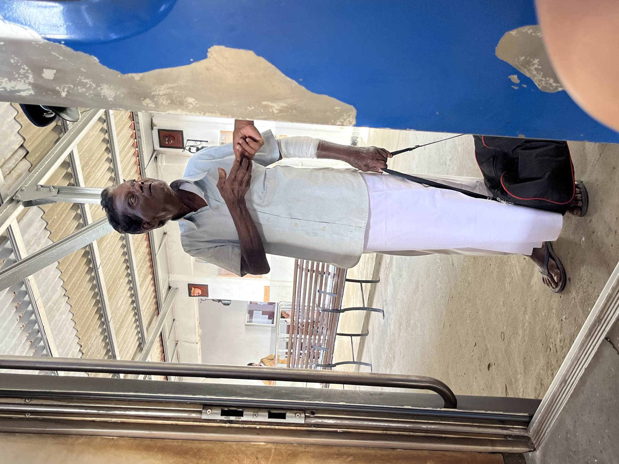 An elderly South Asian man, appearing to be in his 60s or 70s, stands in what appears to be a train station in Ambalangoda, Sri Lanka. He is wearing a light blue collared shirt and a white sarong. His right arm is bandaged from the wrist up to the elbow. He is holding a black duffel bag with red trim by its strap with his left hand. His right hand is reaching out to another person's hand, which is mostly out of frame. The man's expression is one of surprise or concern. The background shows a corrugated metal roof supported by metal beams, and wooden benches typical of a train platform. The floor is concrete. A blue structure with peeling paint is prominently in the foreground on the right.