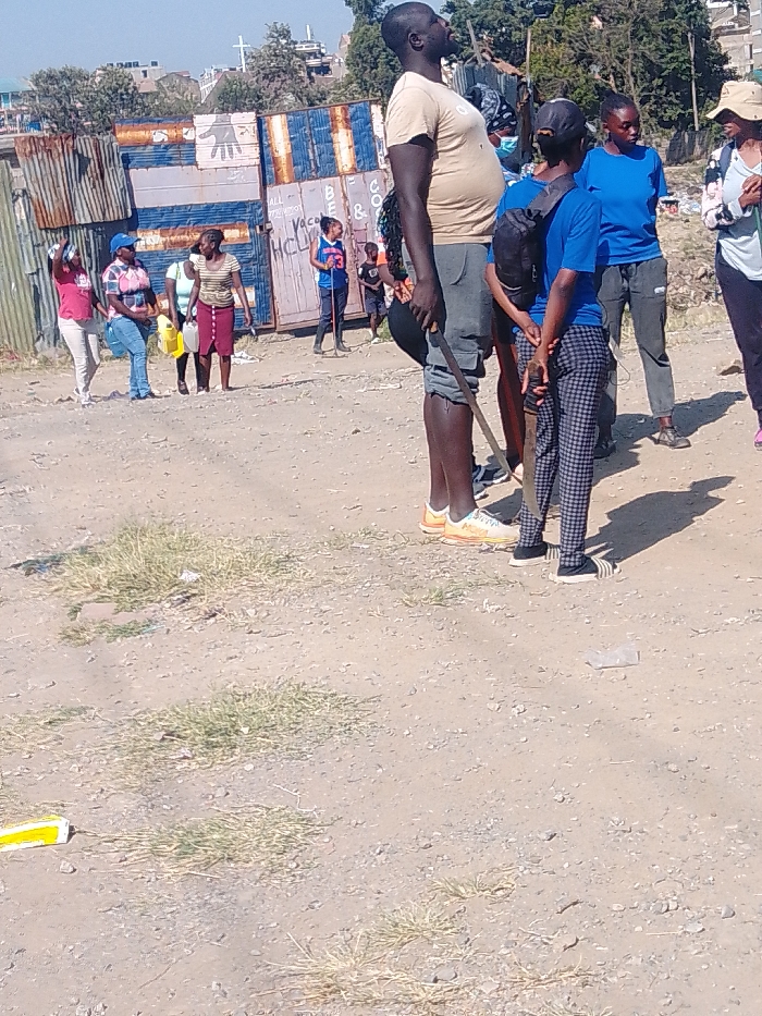 A group of people are gathered on a dirt road in front of shipping containers and corrugated metal fences. One man in the foreground is holding a machete. The people in the background are standing and appear to be waiting. The environment appears to be impoverished.