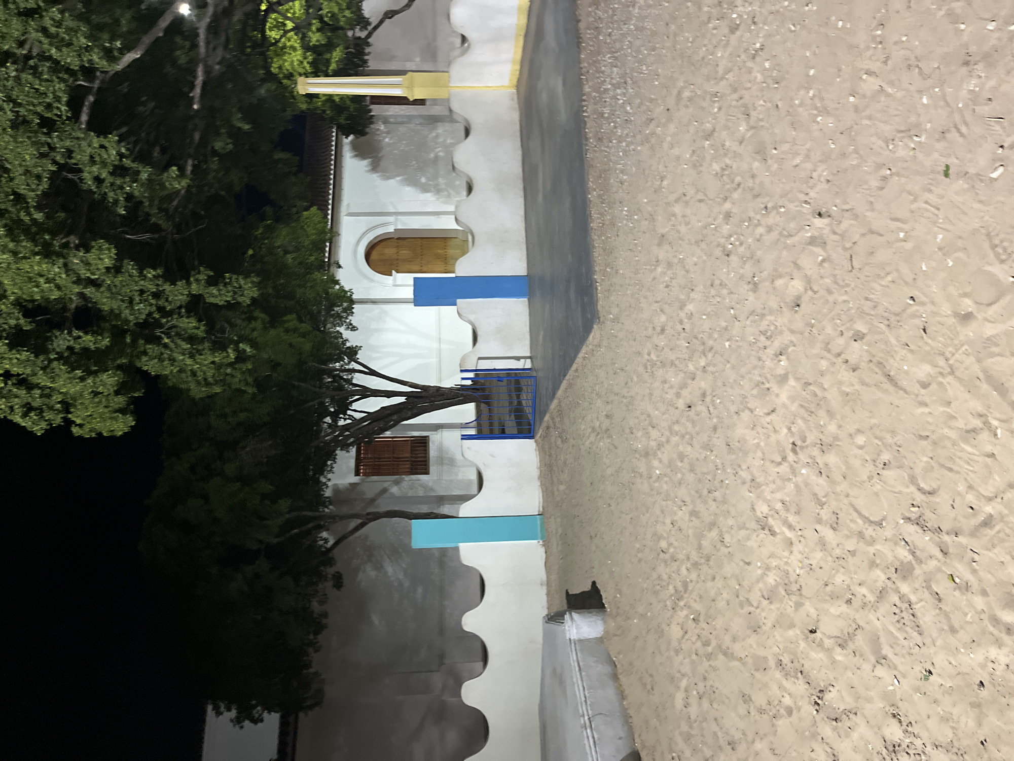 The image depicts an outdoor scene at night, likely in Santa Ana, Venezuela. The foreground is composed of a sandy area with some scattered debris. Behind the sand, there is a low, wavy white wall with curved indentations. To the left of the wall, a cylindrical trash can is visible. Two tall, rectangular pillars, one teal and one blue, stand against the wall.
Beyond the wall is a paved area, partially covered by trees with dark foliage. A building with a white facade and a tiled roof is visible in the background. It features an arched doorway with a wooden door and a small, barred window with wooden shutters. A tall, yellow and white striped pillar is situated to the right of the building. A blue metal gate is present in the middle ground, positioned behind the wavy wall. The overall lighting suggests artificial illumination, possibly from streetlights or building lights, creating shadows and illuminating specific areas of the scene.