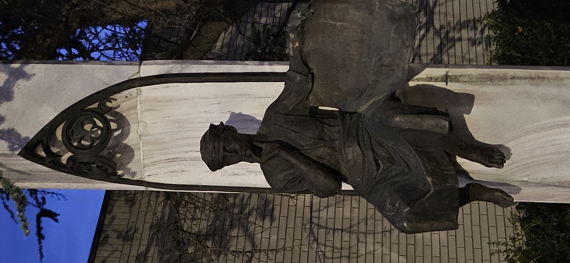 A bronze statue of a pensive figure sits on a pedestal. The figure is dressed in robes and a head covering and is depicted with one hand on its chin, in a posture of contemplation. The statue is situated outdoors against a brick wall. To the right of the statue, a tree with bare branches is visible against a dark blue sky, suggesting either dawn or dusk. The pedestal is made of white stone, with some dark, possibly wrought iron, decorative elements at the top. The statue itself appears to be weathered. This monument is located in the city of Hévíz, Hungary, specifically in front of the former bath attendant's quarters, now part of the Hévíz Museum Collection.