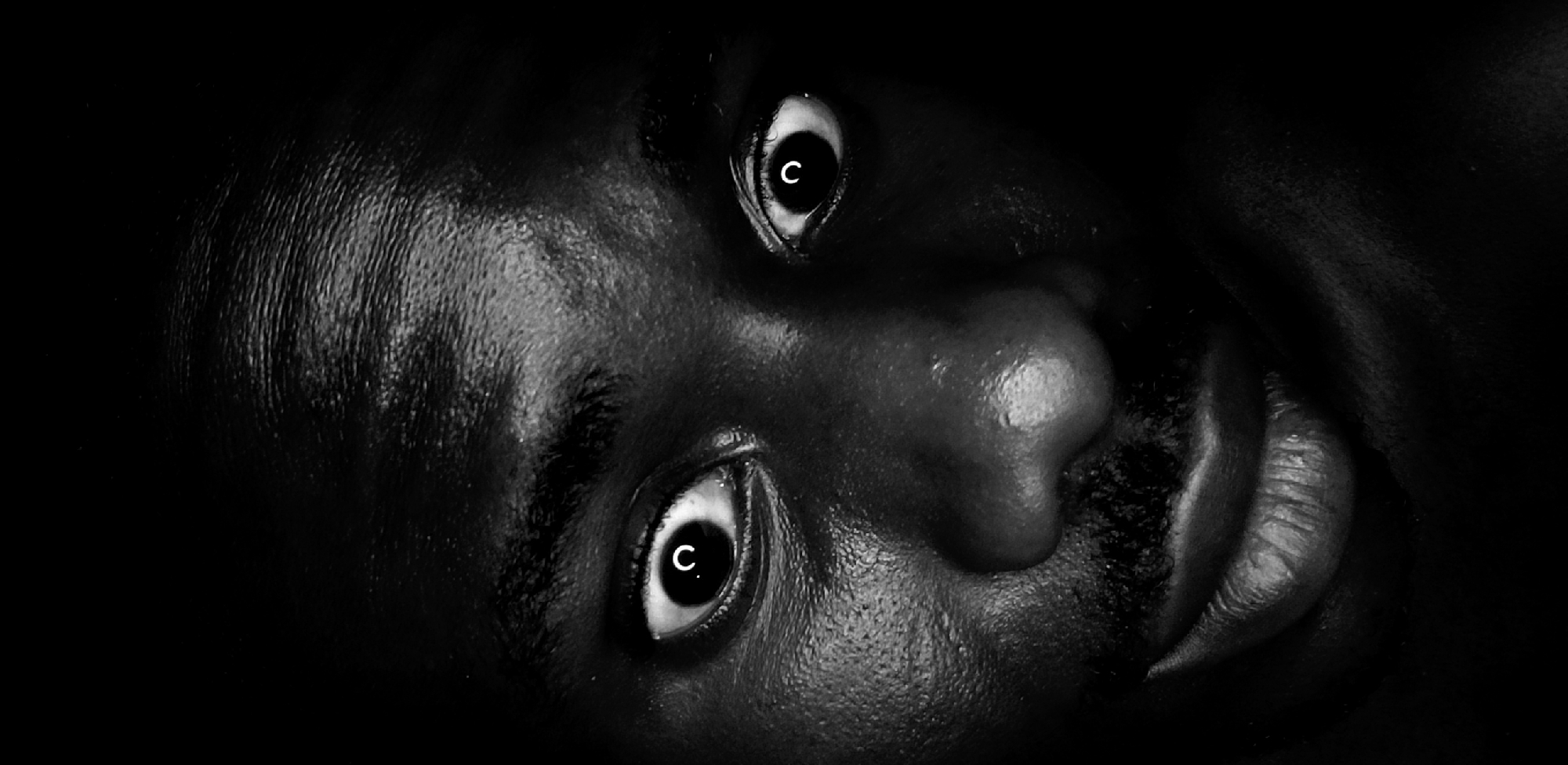 This monochromatic, high-contrast image presents an extreme close-up of a person's face, likely an adult, with dark skin, angled against a pure black background. Dramatic lighting accentuates the contours and textures of the skin on the forehead, nose, and lips, revealing fine lines and pores. The individual's eyes are wide open, with prominent whites and seemingly dilated pupils, each reflecting a distinctive white, capital 'C'. This intense gaze suggests emotions such as alertness, surprise, or perhaps a hint of apprehension or distress. The composition is a static portrait, devoid of any discernible activity or specific scene other than the face itself. The overall aesthetic is stark and intense. This image is from Narayi, Nigeria.