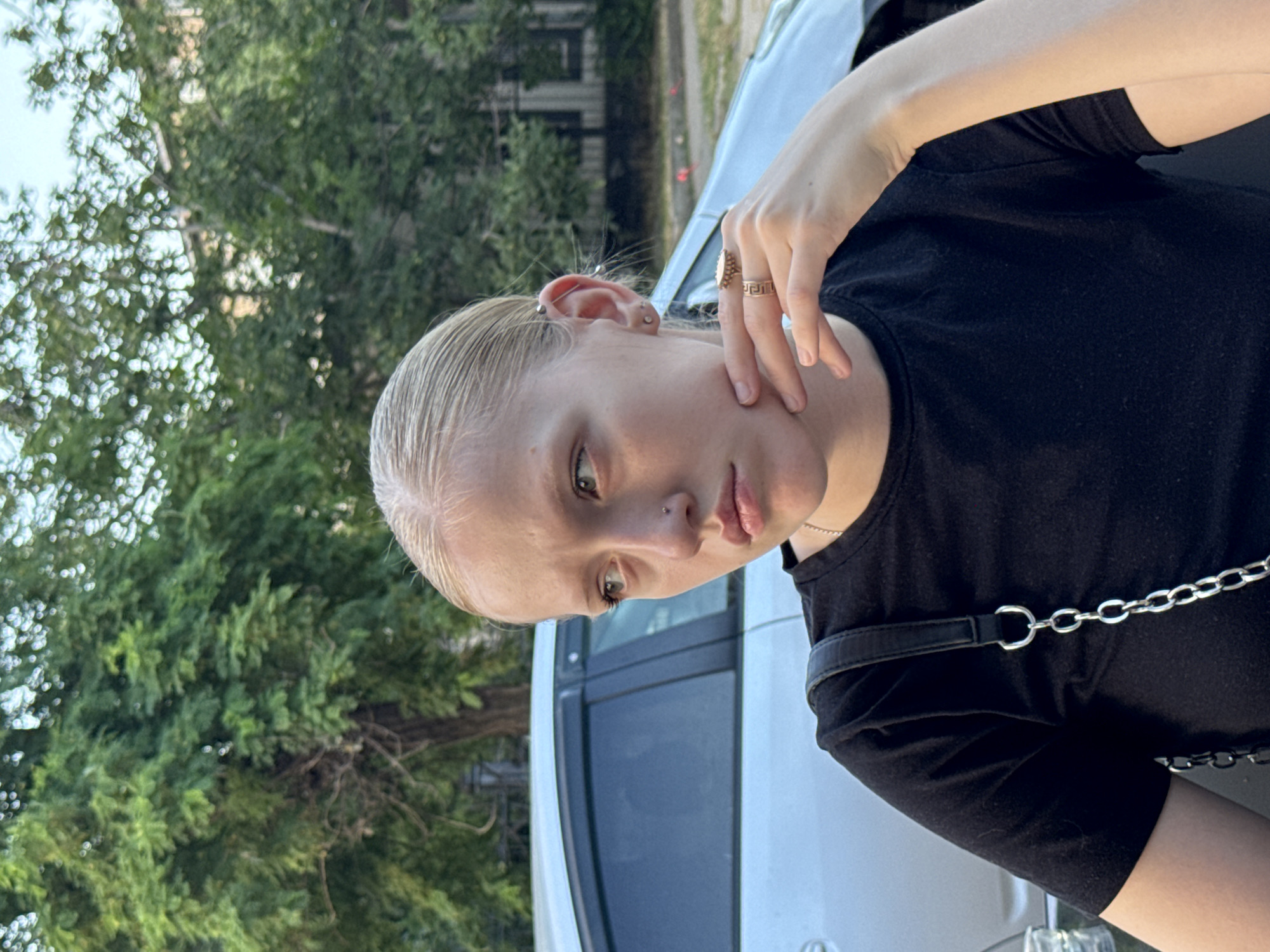A young woman with blonde hair tied back and fair skin is pictured in a close-up, outdoor shot. She is wearing a black t-shirt and has her right hand raised, touching her chin, with rings on two fingers. A silver chain strap from a bag is visible over her left shoulder. She has a piercing on her nose and an industrial piercing on her left ear. The background includes a silver car with tinted windows, lush green trees, and a hint of a building. The lighting suggests it is daytime. The image appears to be taken in the city of Tiraspol, Moldova.