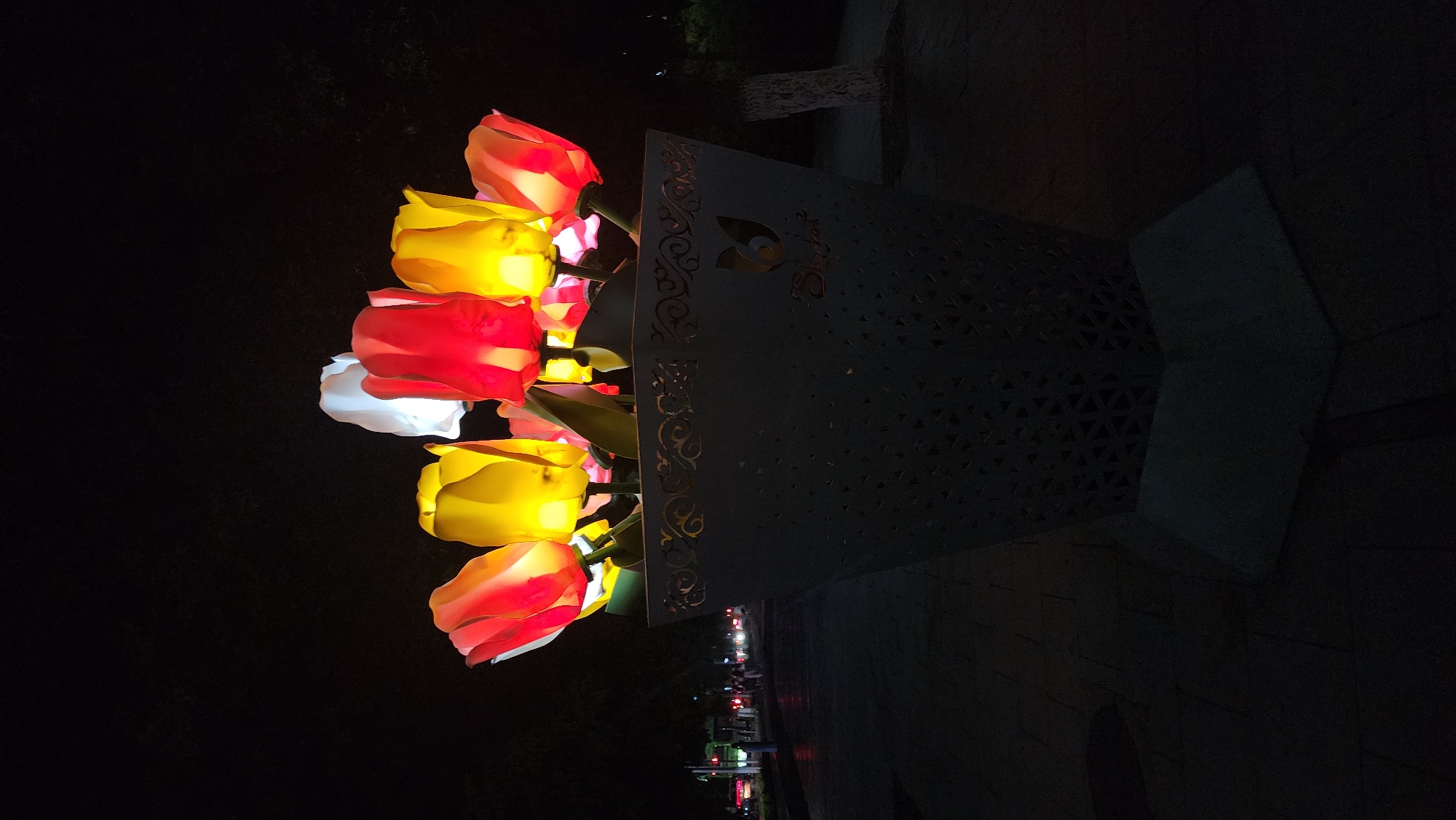 A close-up, low-angle shot features a large, grey, trapezoidal planter filled with illuminated tulip-shaped lanterns against a dark, night sky. The lanterns are predominantly red and yellow, with one white lantern visible towards the back. The planter has a decorative, laser-cut geometric pattern near the top and a logo that appears to read "Shymkent" beneath a stylized tulip. The planter sits on a paved walkway, with a blurred background showing a street with traffic lights and some distant pedestrians. The overall impression is of an outdoor decorative installation in the city of Shymkent, Kazakhstan, likely along a pedestrian area or street.