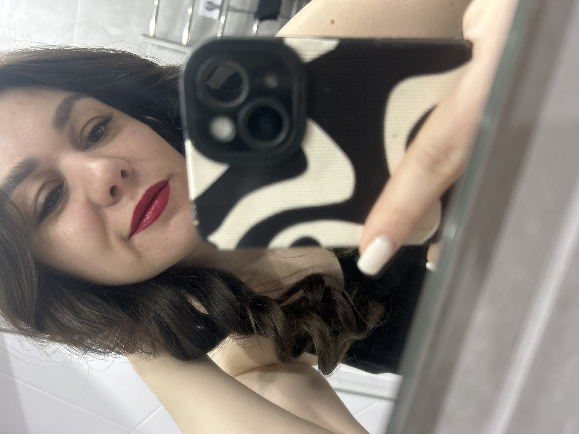 A woman with dark hair and red lipstick takes a selfie in a bathroom. She is holding a phone with a black and white patterned case. The bathroom has white tiles and a metal towel rack. The location is Chaikovsky, Russia.
