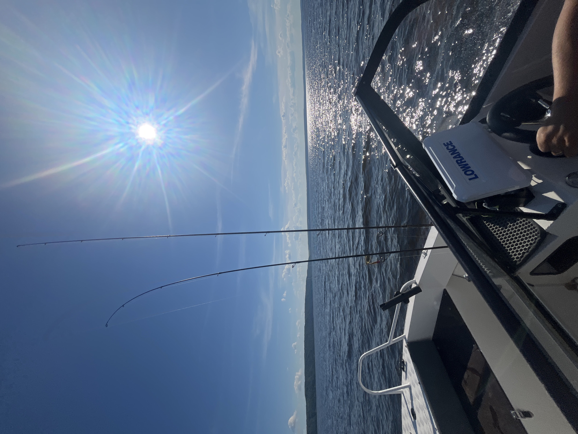 This media file depicts a daytime scene from a boat on a body of water, likely during a fishing trip. The sun is shining brightly, creating a strong lens flare across the upper left portion of the image and reflecting off the water's surface. The sky is a clear, bright blue with some scattered white clouds.
The main subjects are fishing rods positioned in rod holders on the boat. Two fishing rods are clearly visible, extending upwards and out over the water. One rod is bent, suggesting it might be in use or has a weight attached. A fishing reel is visible on one of the rods.
The boat itself is partially shown, with the console and part of the seating area visible on the right side of the frame. A person's hand is seen on the steering wheel, indicating they are piloting the boat. A "LOWRANCE" branded fishfinder or GPS unit is prominently displayed on the console. The water is calm with ripples, reflecting the sunlight. In the distance, a hazy landmass or shoreline is visible along the horizon.
The overall mood is one of leisure and outdoor activity, specifically fishing. The bright sun suggests it is midday. The presence of fishing equipment and the boat indicates a recreational activity taking place on a lake, river, or sea. The visible text "LOWRANCE" is a brand name for marine electronics. Given the provided location context of Siukterka, Russia, this scene likely takes place on one of the local waterways in that region.