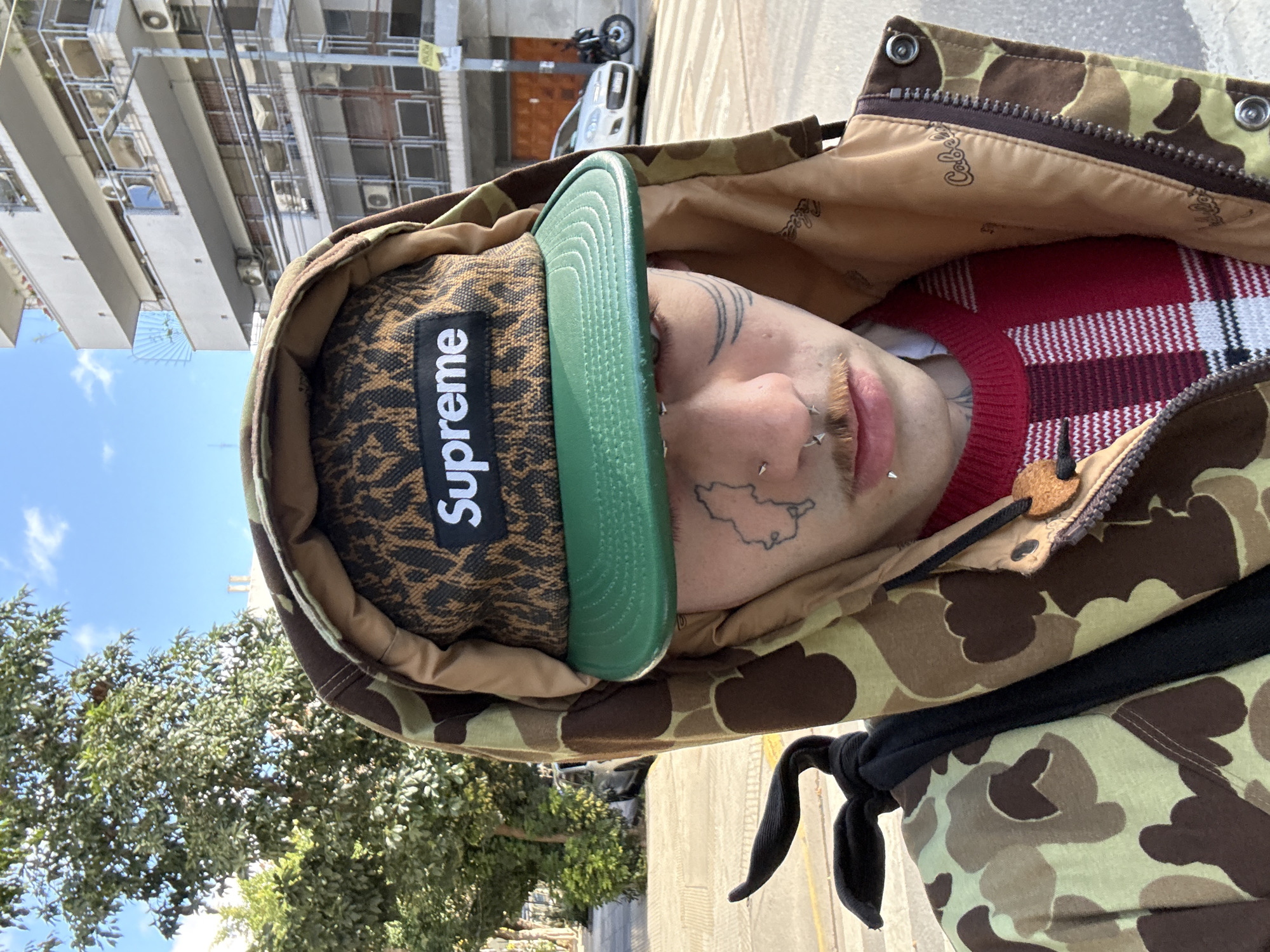 A person wearing a Supreme hat and a camouflage jacket is outdoors in Buenos Aires, Argentina. The hat is brown with a black "Supreme" logo, and a green brim. The jacket is camouflage with a brown and tan pattern. The person has tattoos on their face and piercings on their eyebrows and nose. A red and white checkered garment is partially visible under the jacket. In the background are a building with multiple windows, a blue sky, trees, a parked car, and a motorbike. A black drawstring is tied around the person's jacket.
