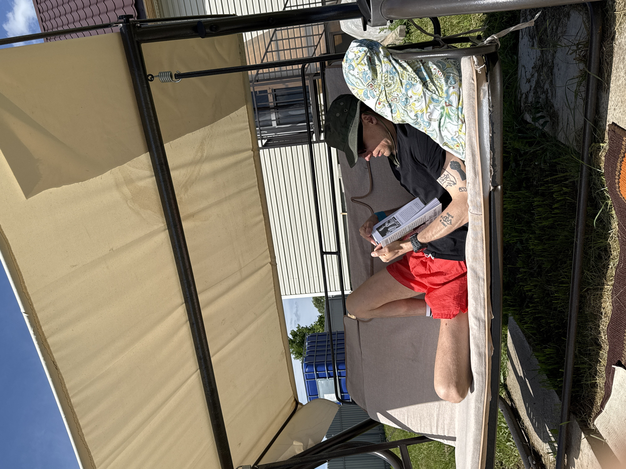 A man with tattoos on his arm is sitting on a swing set, reading a book. He is wearing a hat, sunglasses, a black t-shirt, and red shorts. He is sitting on a cushion with a patterned pillow behind him. The swing set is outdoors, with a tan canopy overhead. In the background, there is a house with a fence and some large blue tanks. The scene appears to be in a suburban area, possibly the city of Serpukhov, Russia.