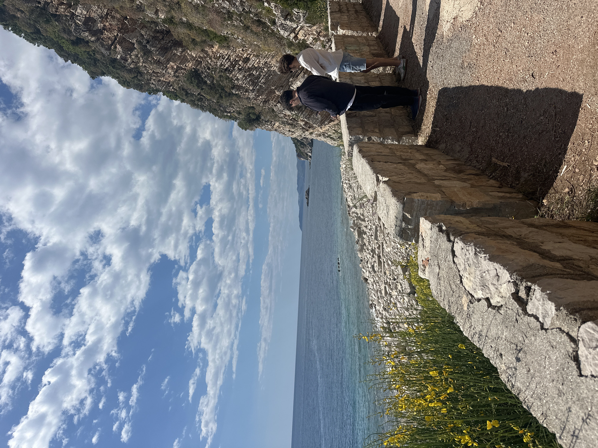 The image shows a coastal scene in Perazića Do, Montenegro. Two people are positioned on a stone pathway or ledge, overlooking the Adriatic Sea. One person is wearing a dark jacket and the other a light-colored shirt. The landscape includes a rocky mountain ridge on the left side, a blue sky with scattered clouds, and the sea. Yellow flowers grow along the edge of the pathway. The composition gives a sense of height and the viewpoint offers a view along the Montenegrin coast.