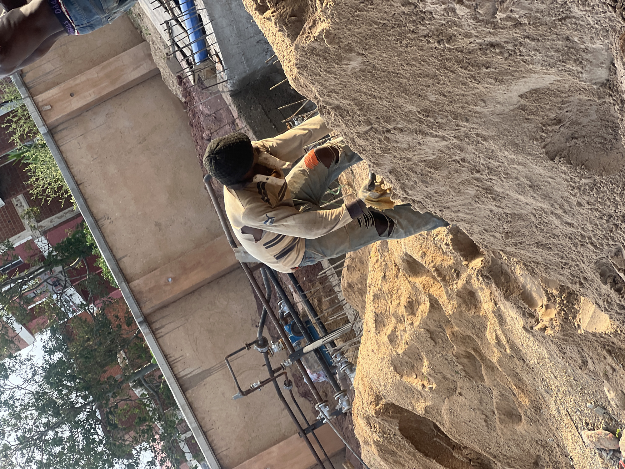This image captures an active outdoor construction or renovation site in Jos, Nigeria, under clear, sunny skies, likely in the late morning or afternoon.
In the mid-ground, a man is engaged in manual labor, his back largely to the viewer. He wears a light-colored, long-sleeved shirt, work trousers (possibly denim jeans) with a prominent rip on the right knee revealing an orange-red layer beneath, and a dark cap or head covering. A work glove is visible on his left hand. His posture suggests he is bending over, possibly manipulating elements of the nearby pipe system or handling materials.
To the right of the worker, a large, textured mound of sand or dirt dominates the frame. Directly behind the man, an intricate network of metal pipes and valves, along with exposed rebar, indicates ongoing plumbing and structural work. The ground around these elements consists of disturbed earth and loose sand.
In the upper left of the image, the lower torso and legs of another individual are visible, wearing blue denim shorts. The elastic waistband of their underwear or inner garment is exposed, bearing text that appears to read "HIPPO SPORT" or "SITOPPO".
The background on the left side features green foliage from trees and bushes, partially obscuring brick buildings with red roofs, offering a glimpse of the surrounding residential or urban landscape. Metal beams or structural elements are also visible, reinforcing the construction setting. The scene conveys a sense of diligent labor in an active work environment.