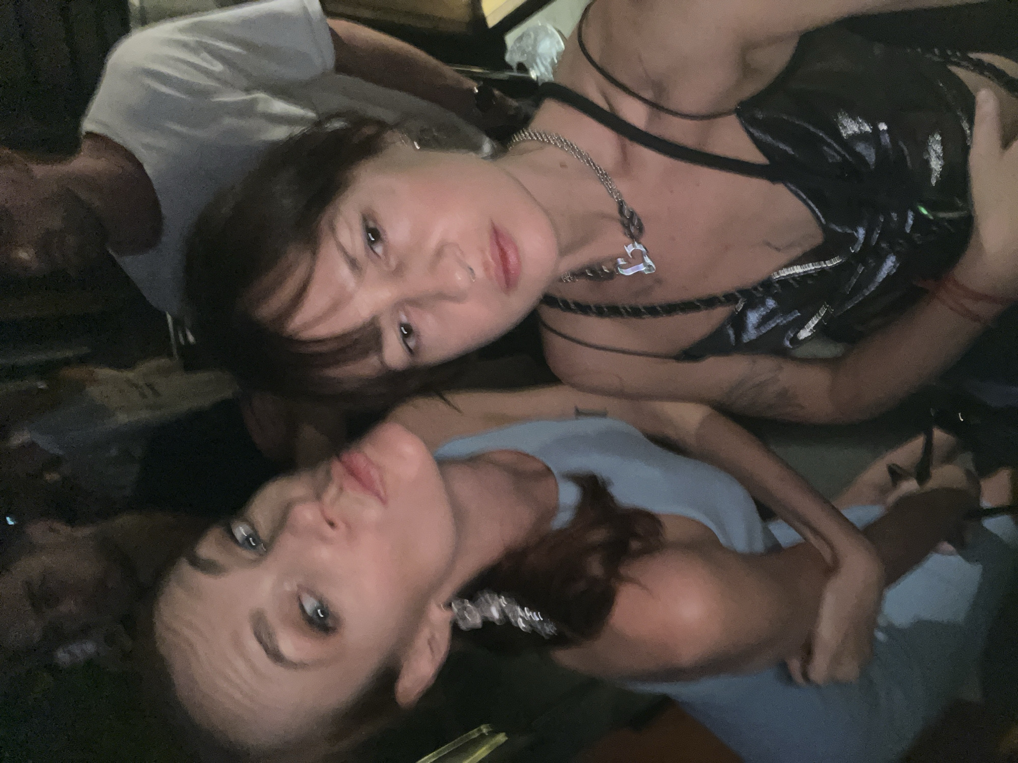 The image shows two women posing for a photo in a dimly lit, intimate setting, likely at night in North Kuta, Indonesia. The camera angle is from slightly above, with the subjects appearing to lie down or lean back.
The woman at the top of the frame has dark hair and wears a dark, possibly patent leather or vinyl, top with thin straps. She accessorizes with a silver chain necklace featuring a heart-shaped pendant and has a subtle nose piercing on her left nostril. Her expression is direct and neutral, looking towards the camera.
Below her, another woman with lighter brown hair is visible. She wears a light blue top and a silver-toned, possibly chunky, necklace or choker. Her lips are slightly pursed in a subtle pout as she also looks towards the camera.
Portions of other individuals are discernible in the background; a person in a white t-shirt is partially visible behind the top woman, and another figure is dimly seen behind the bottom woman. The background itself is dark and indistinct, indicating artificial lighting conditions. No text is visible in the image. The overall ambiance suggests a social gathering.