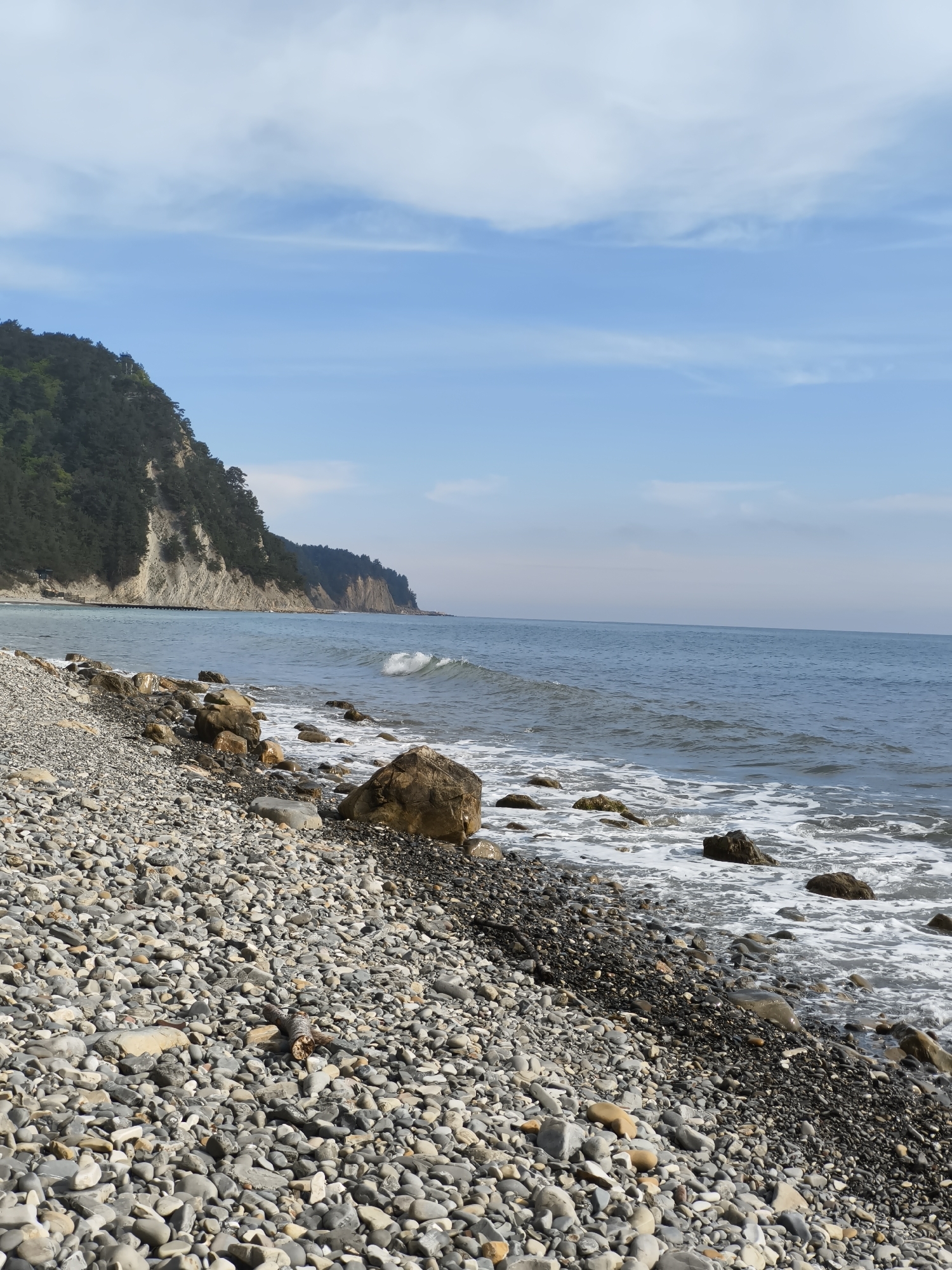 The image depicts a pebble beach in Praskoveyevka, Russia. The shoreline consists of small, rounded grey and black pebbles interspersed with larger rocks. Waves are breaking along the shore, creating white foam. To the left of the frame, a hill covered in dark green vegetation rises sharply from the beach. The sky is a pale blue with a few scattered white clouds. The sea is a dark blue-grey, stretching to the horizon. There are no people visible in the scene.