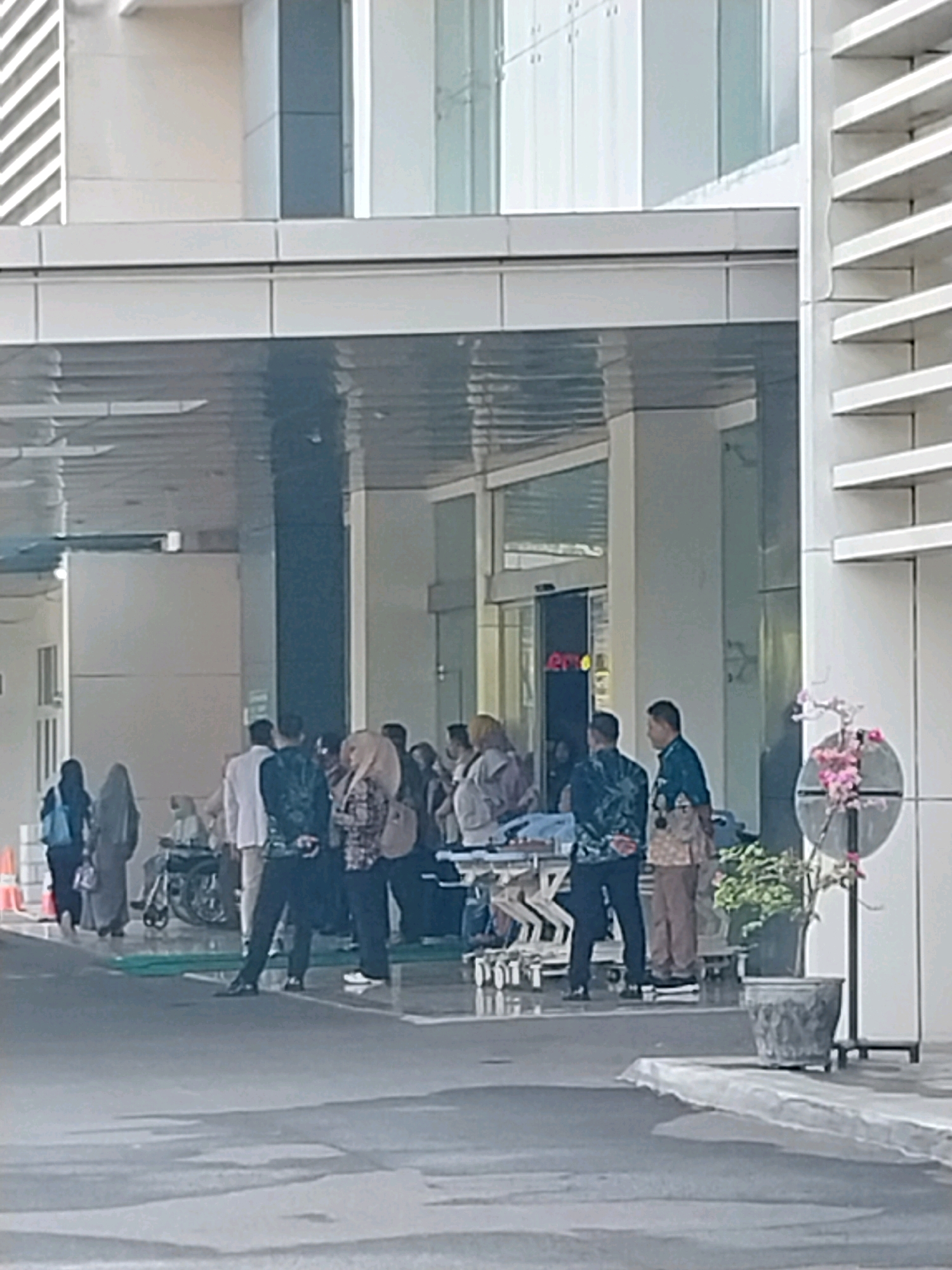 The image depicts the exterior entrance of a modern building, likely a hospital, in Wates, Indonesia. A group of people are gathered under a covered walkway, some in conversation and others appearing to be attending to a person on a gurney. The architecture features clean lines, glass doors, and large windows. A prominent feature is a decorative floral arrangement in a planter to the right of the entrance. The ground is paved, suggesting an outdoor area such as a driveway or plaza.