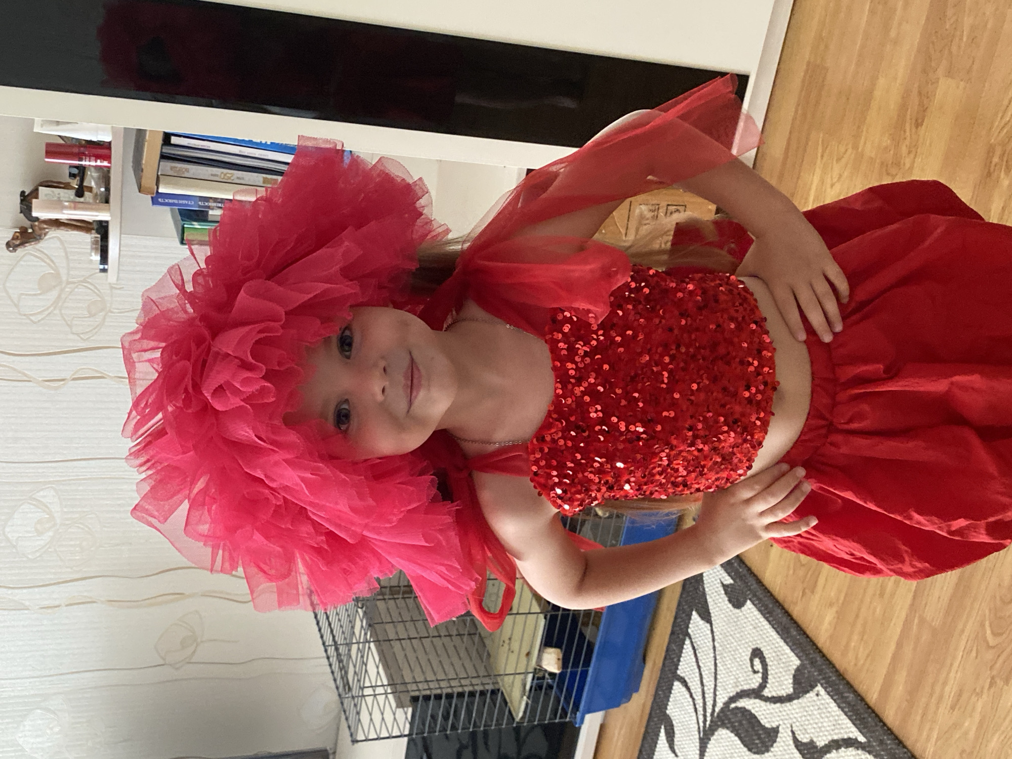 A young girl with blonde hair and blue eyes, dressed in a sparkly red crop top and a red skirt, poses for a photo in a room. She wears a large, fluffy pink tulle headdress and a red sheer fabric draped around her shoulders. The girl is positioned in the center of the frame, with her left hand resting on her stomach and her right hand on her skirt. She is smiling at the camera.
In the background, a bookshelf with various books and cosmetic items is visible. To the right of the girl, a wooden floor and a rug with a black and white floral pattern are present. A black metal cage and a blue plastic container are also in the background, suggesting a home environment. The lighting appears to be indoor, with no clear indication of the time of day or weather. The overall mood of the image is cheerful and playful, likely taken during a dress-up or performance moment. No visible text is present in the image.