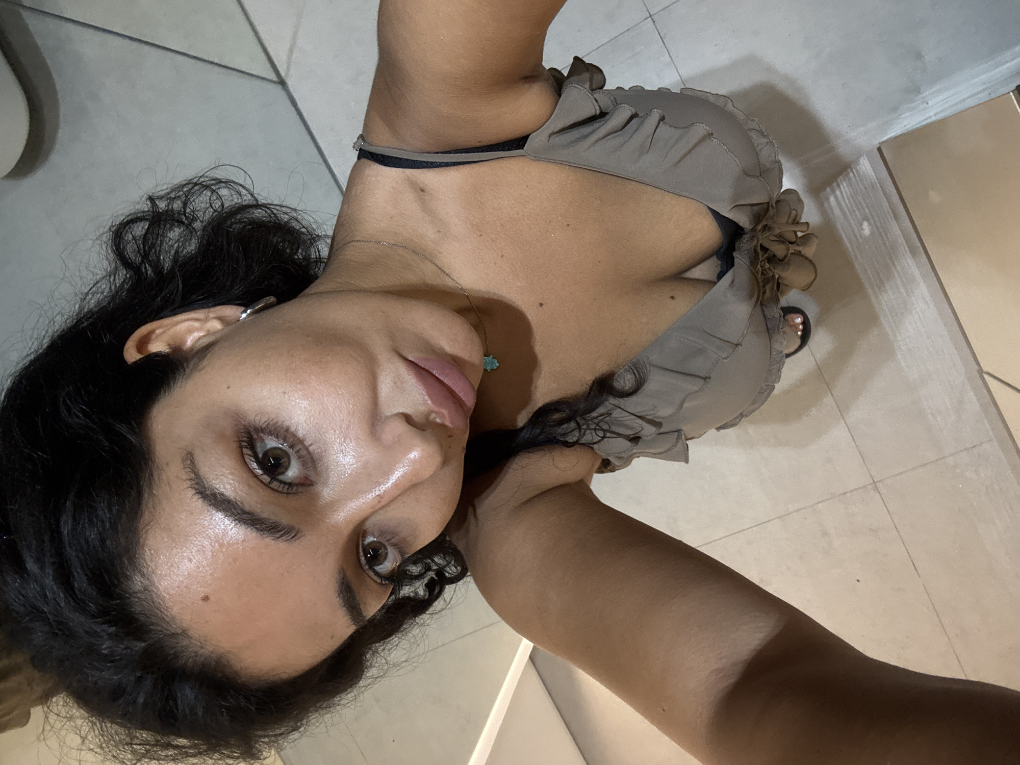 A close-up selfie shows a woman looking directly at the camera with a neutral expression. She has dark, curly hair framing her face and flowing down her back. Her large brown eyes are prominent, accentuated by visible mascara and eyeliner. Her eyebrows are dark and well-defined. Her skin has a healthy glow, particularly on her forehead and cheeks. She is wearing a light gray or beige top featuring ruffled details on the shoulders and neckline, with a dark bra strap visible. Around her neck, she wears a silver-colored chain necklace adorned with a small, light blue Hamsa hand pendant. A silver-colored hoop or cuff earring is visible in her left ear. The background primarily consists of light-colored tiled flooring and a plain wall or fixture, suggesting an indoor setting. Her legs and a black sandal on her right foot are partially visible in the lower part of the frame. No text is visible in the image. The provided context indicates the location is Kuta Selatan, Indonesia, though the indoor setting does not offer specific geographical cues.