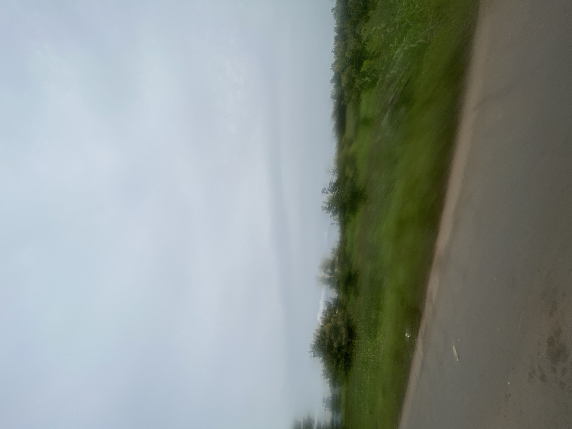 The image shows a blurred landscape with a grey sky overhead. In the foreground, there is a dark asphalt road extending diagonally from the bottom left to the middle right of the frame. To the right of the road, a grassy field is visible, slanting upwards and covered with green vegetation. Scattered across the field are blurry green trees and bushes. The overall impression is of a motion-blurred scene captured from a moving vehicle. The location is specified as Gidan Waya, Nigeria, suggesting a rural or semi-rural setting with natural greenery and a road. There are no people or specific objects clearly identifiable due to the motion blur.