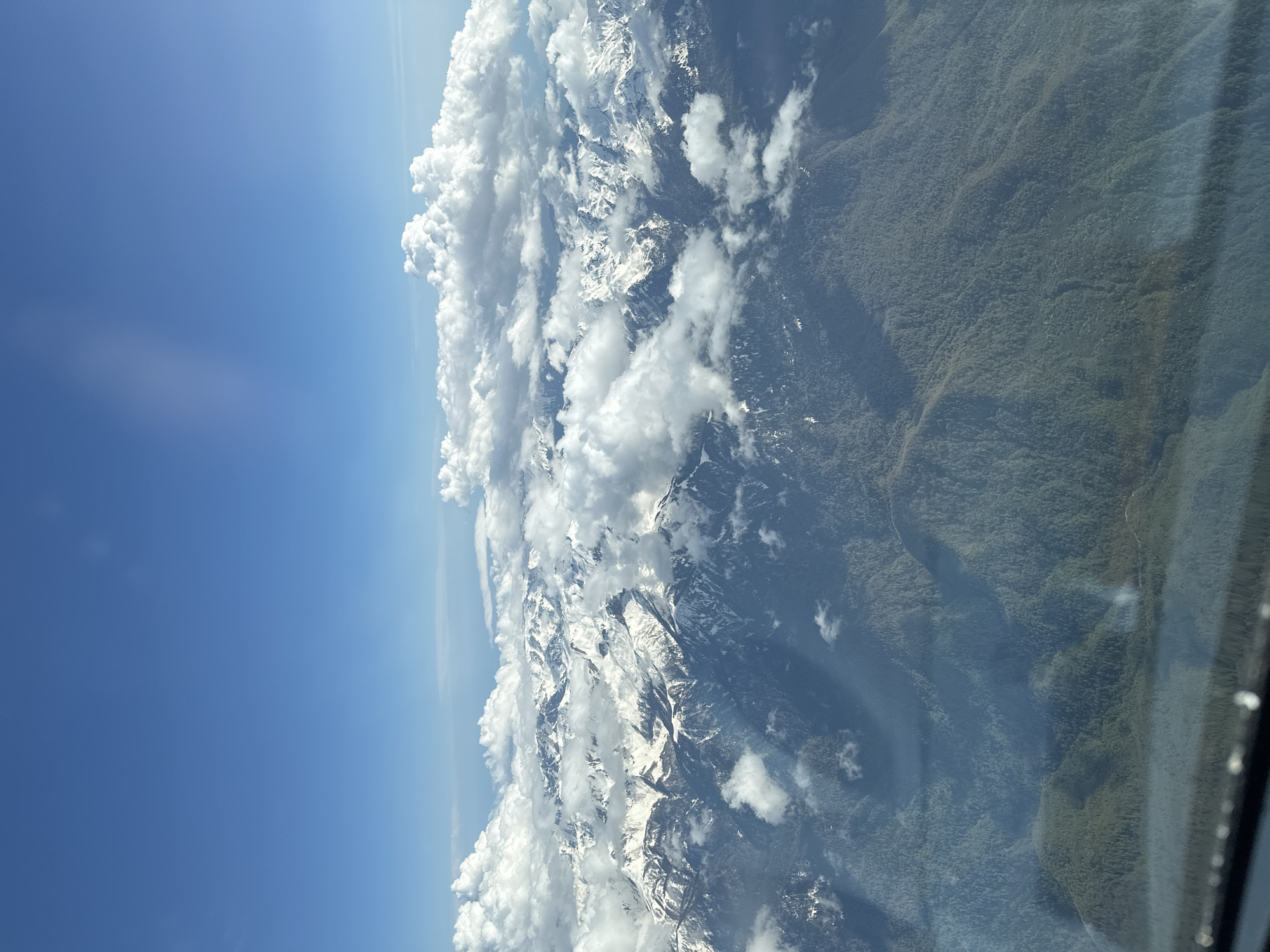 The image captures a mountainous landscape in Sochi, Russia, viewed from an elevated position, possibly an airplane window. Snow-capped mountains intermingle with puffy white clouds under a clear blue sky. Dense forest covers the lower slopes of the mountains. A part of a window frame is visible on the right side of the image.