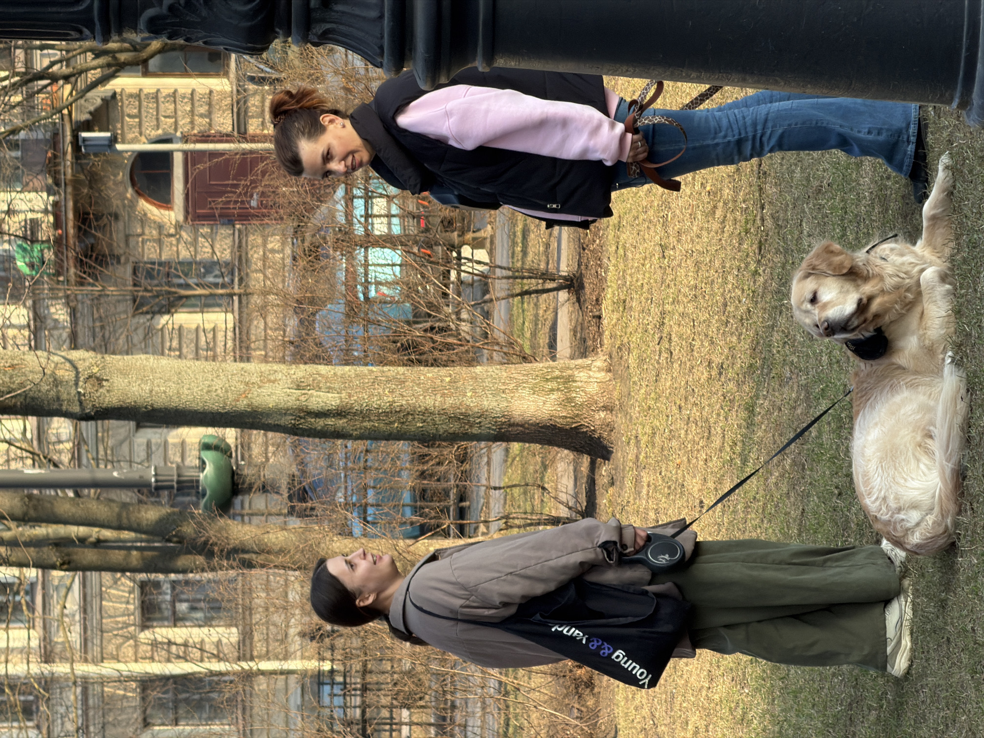 The image depicts two women and a golden retriever dog in a park setting in Saint Petersburg, Russia. One woman, wearing a pink sweatshirt, black vest, and jeans, stands near a black lamppost, looking down at the dog. The dog is lying on the grass. The other woman is walking the dog, wearing green trousers and a jacket. In the background, the architectural details of Saint Petersburg buildings are visible, with bare trees filling the middle ground.
