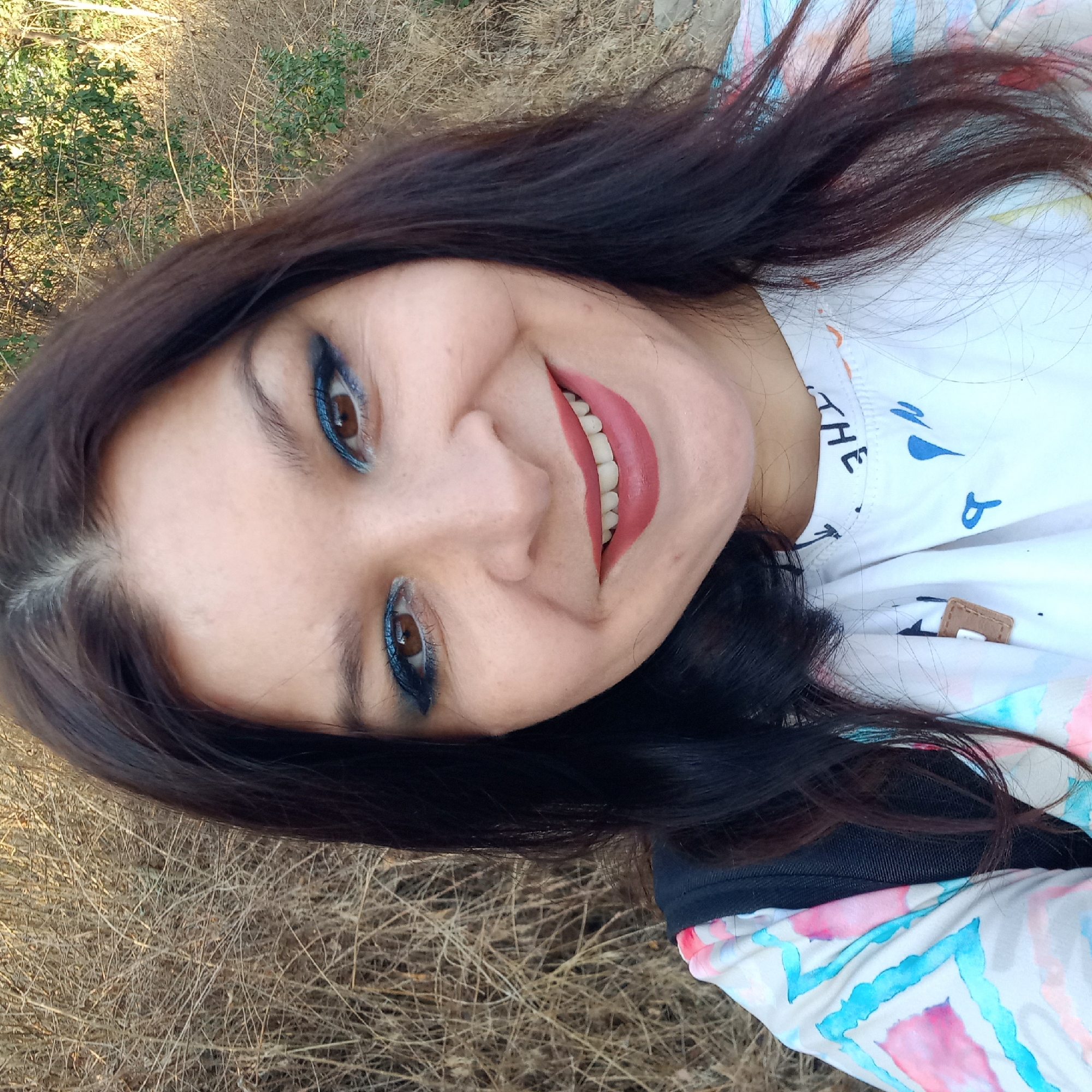 A woman with dark hair and blue eyeshadow smiles at the camera. She is wearing a white t-shirt with a colorful, abstract pattern of pink, blue, and white shapes. A black backpack strap is visible over her shoulder. The background consists of dry, brown vegetation and some green foliage, suggesting an outdoor setting. The lighting indicates it is daytime. The image is a selfie taken from the chest up. The specific location in Klyntsi, Ukraine, cannot be determined from the visual information provided.