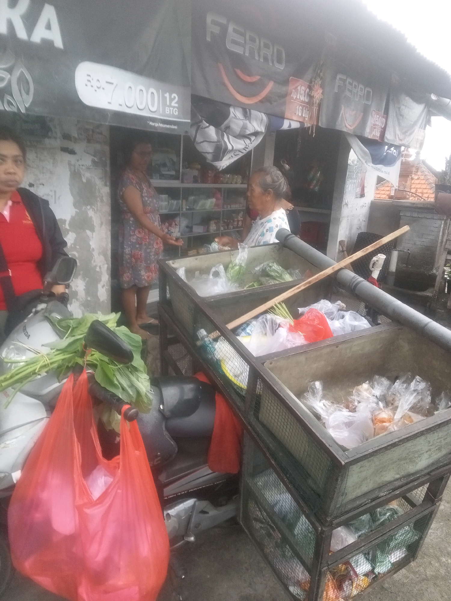 The image depicts an outdoor stall or shop in what appears to be the city of Mendoyo, Indonesia. The stall has a corrugated metal roof with banners displaying "FERRO" and product pricing. A prominent sign reads "Rp. 7.000 12 BTG". Two women are visible: one on the left wearing a red shirt and black jacket, standing next to a silver scooter with a red plastic bag and green vegetables hanging from the handlebars. The other woman, on the right, wears a floral dress and is standing in front of shelves stocked with goods. In front of the stall, there are two stacked metal cages filled with various items, many of which are packaged in plastic bags. One cage is open at the top, containing more plastic-wrapped items. The ground appears to be concrete or asphalt. The overall scene suggests a marketplace or a small retail establishment.
