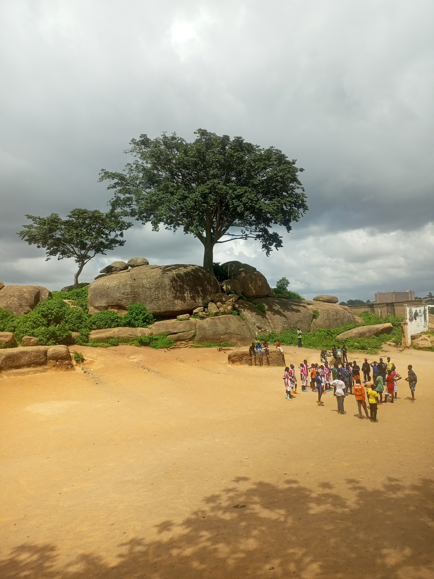 The image depicts a natural rock formation in Jos, Nigeria, with a group of people gathered in the foreground. The rock formation is characterized by large, weathered boulders interspersed with green vegetation and trees. The sky is overcast with dramatic clouds, suggesting a potential for rain.
In the foreground, a sandy, open area serves as the gathering point. A group of approximately 25 to 30 individuals, predominantly young males, are clustered together. Many are wearing athletic attire, including jerseys in various colors like red, white, yellow, and orange, suggesting a sports-related activity or a casual congregation. Some individuals are observing the group, while others appear to be engaged in conversation or waiting.
To the right of the rock formation, a partially visible building with multiple stories stands, indicating the presence of urban development in the vicinity. A low wall, possibly a boundary, separates the natural area from the constructed environment. A portion of a mural or signage is visible on the wall. The ground is a mixture of sand and dirt, with shadows of trees cast upon it, indicating the time of day. The overall scene captures a moment of human activity within a natural landscape, set against the backdrop of a cloudy sky and distant urban structures in Jos, Nigeria.