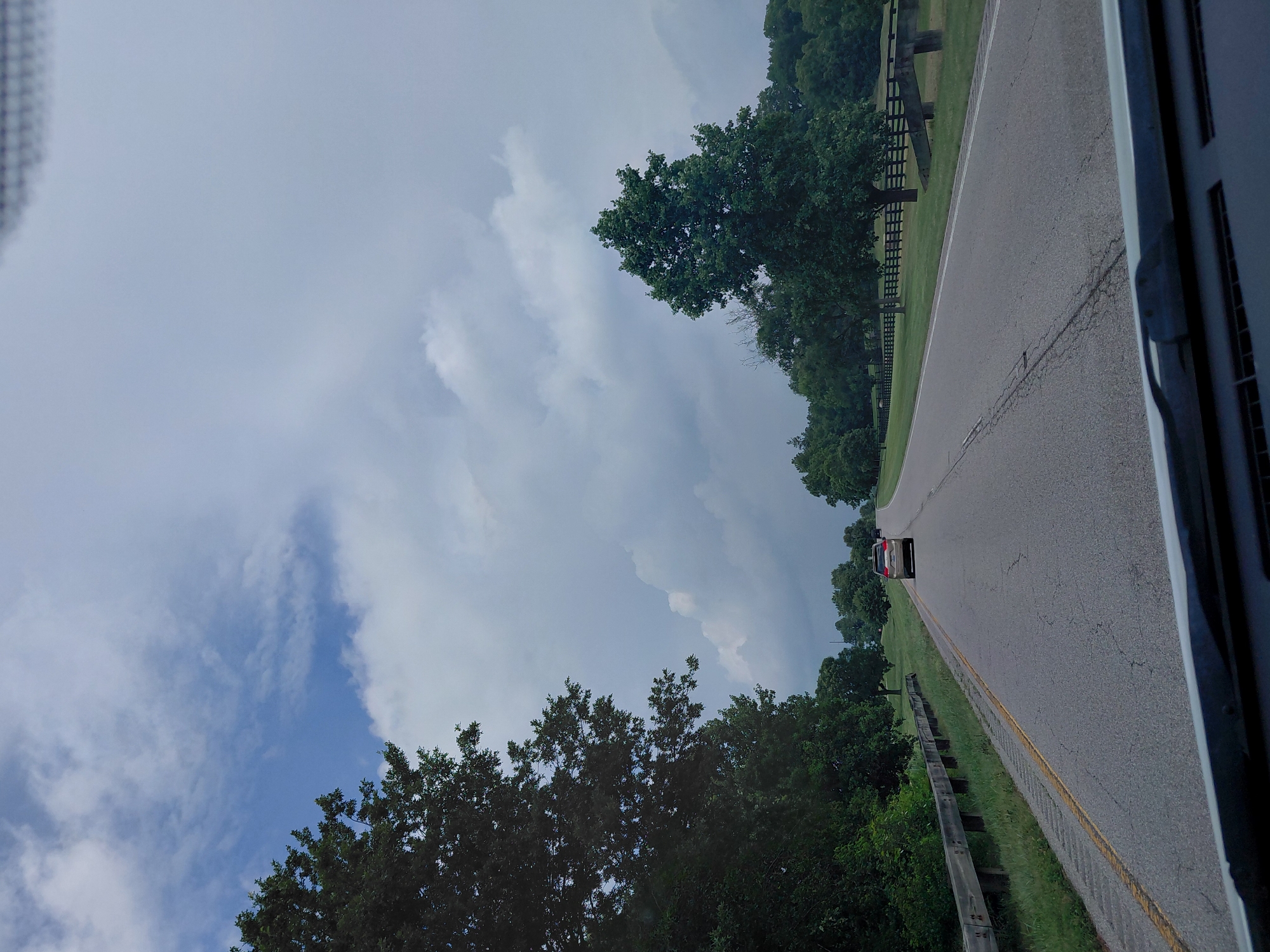 A vehicle drives down a two-lane asphalt road on a cloudy day in Paris, United States of America. The road is bordered by a guardrail on the left and trees and a black wooden fence on the right. In the distance, a few more vehicles are visible. The sky is filled with dramatic clouds, suggesting an approaching storm. The overall scene depicts a rural or semi-rural drive through a wooded area.