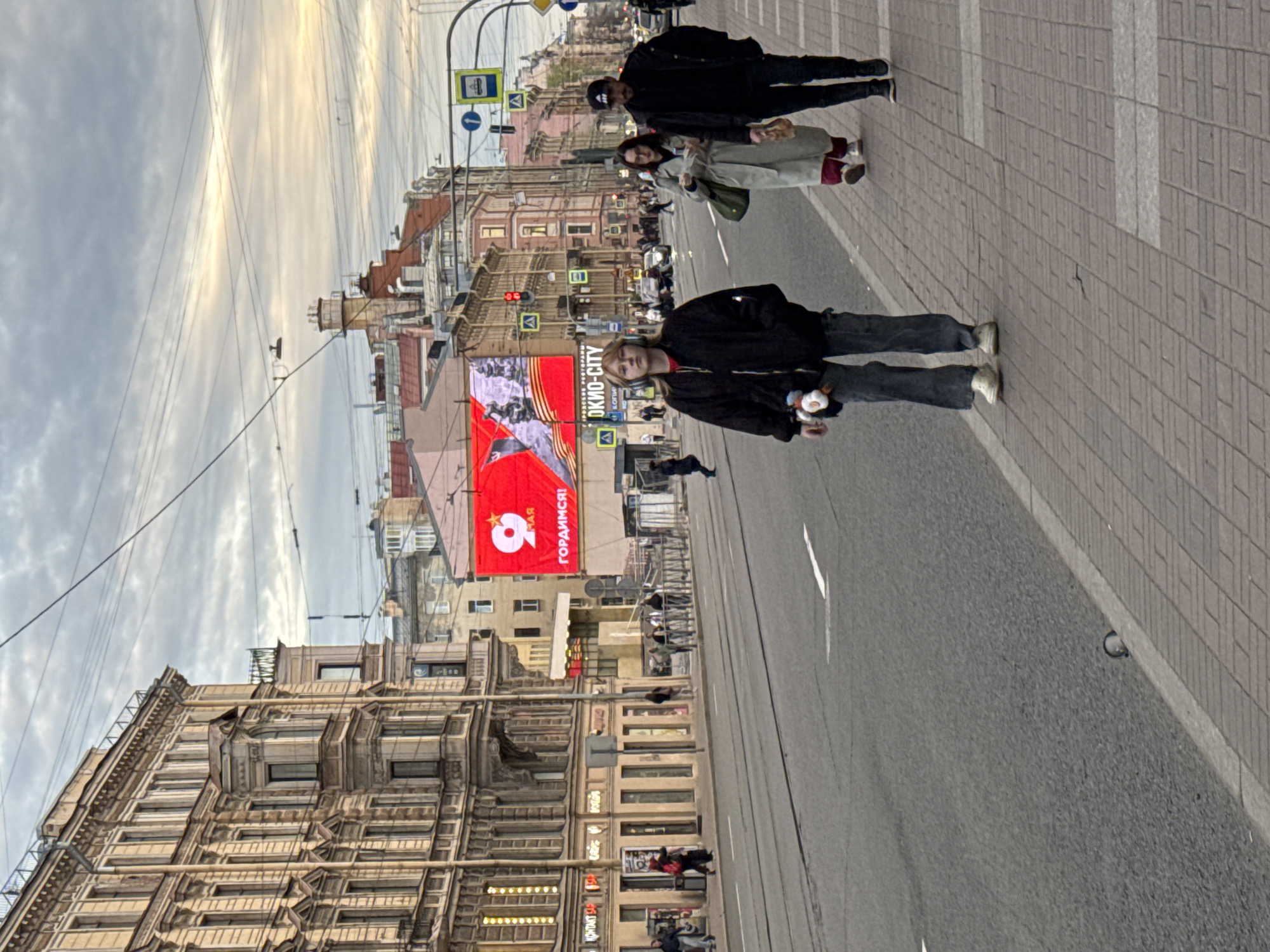 The image shows a street scene in Saint Petersburg, Russia. Several people are walking on the sidewalk. The architecture consists of old, ornate buildings. Overhead wires run above the street. A large banner with the number "9" and Cyrillic text hangs on one of the buildings. Traffic signals are visible along the street.