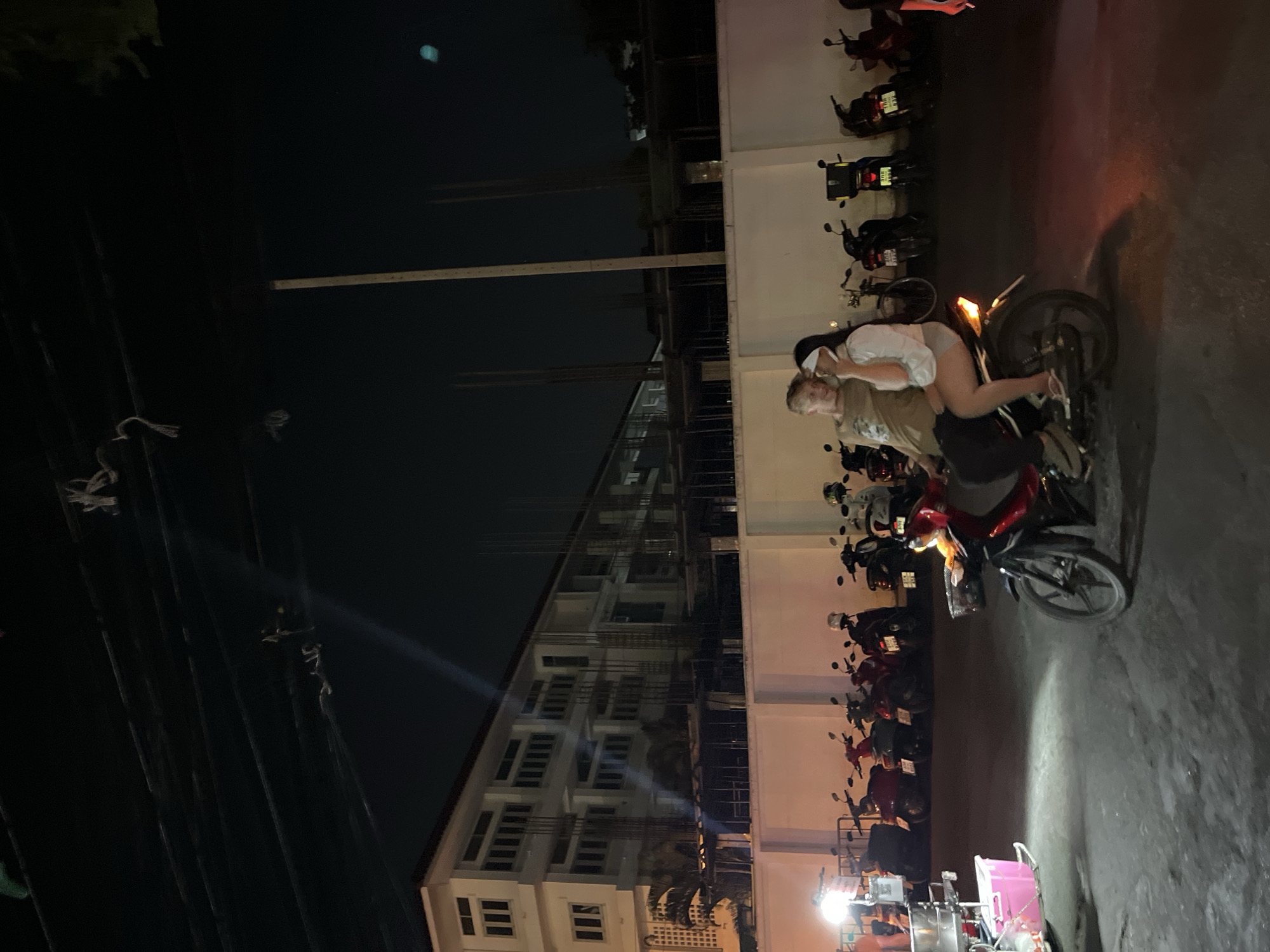 A nighttime urban scene in Chiang Mai City Municipality, Thailand. Two people, one fair-skinned male wearing a khaki shirt and one fair-skinned female wearing a white shirt and grey shorts, are sitting on a red scooter. The male is seated in front while the female is behind him, with her arms around his waist. The scooter's taillight is illuminated. They are parked along a street lined with parked scooters, arranged in rows against a tall, white building with multiple floors and windows. A building under construction is adjacent. Power lines are visible overhead, and a streetlight illuminates a vendor.