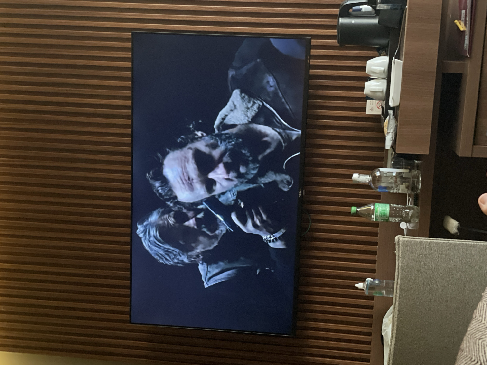 The image shows a television mounted on a horizontally ribbed wooden wall. The television screen displays a scene with two men, one older with gray hair and another younger man, both in what appears to be a dramatic scene. To the right of the television is a dark wooden cabinet. It holds items such as a black electric kettle, white cups, various packets, and bottles of liquid. A gray upholstered headboard of a bed and a person's hand are visible in the lower right corner of the frame. The image was taken indoors. There is no information to ascertain if this photo was taken in Otopeni, Romania.