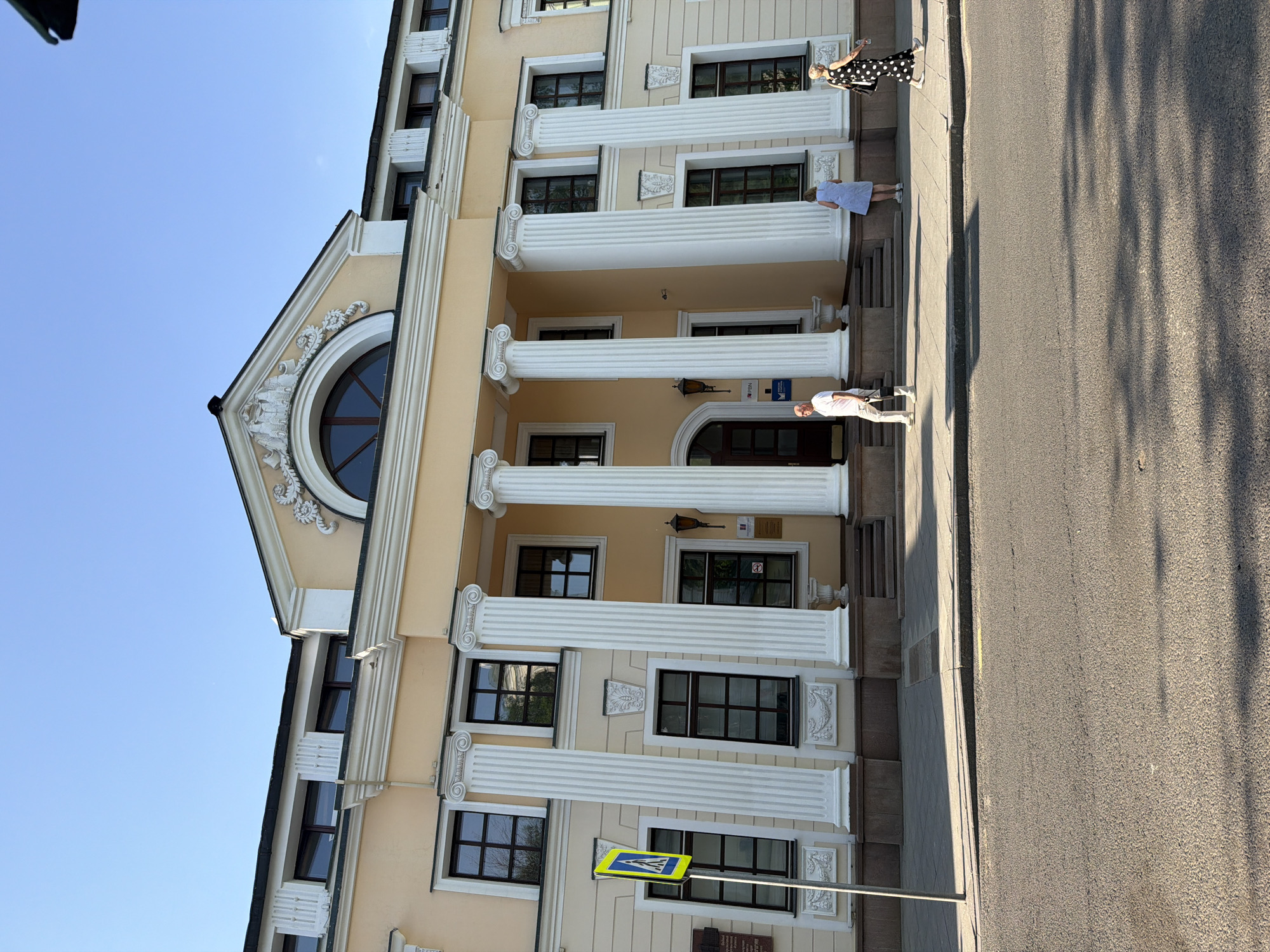 The image shows a light yellow building with white columns located on a street in Moscow, Russia. The building has a triangular pediment with a round window and ornate detailing. The facade has multiple windows with dark frames. There are three people on the sidewalk in front of the building. The first, dressed in a white shirt and pants, stands on the steps. The second person, in a light blue dress, stands a few steps above the first. The third person, dressed in a black and white polka dot dress, stands closest to the top of the steps. A yellow pedestrian crossing sign is planted in the ground in front of the building. A paved road runs along the sidewalk's edge. Tree shadows stretch across the road.