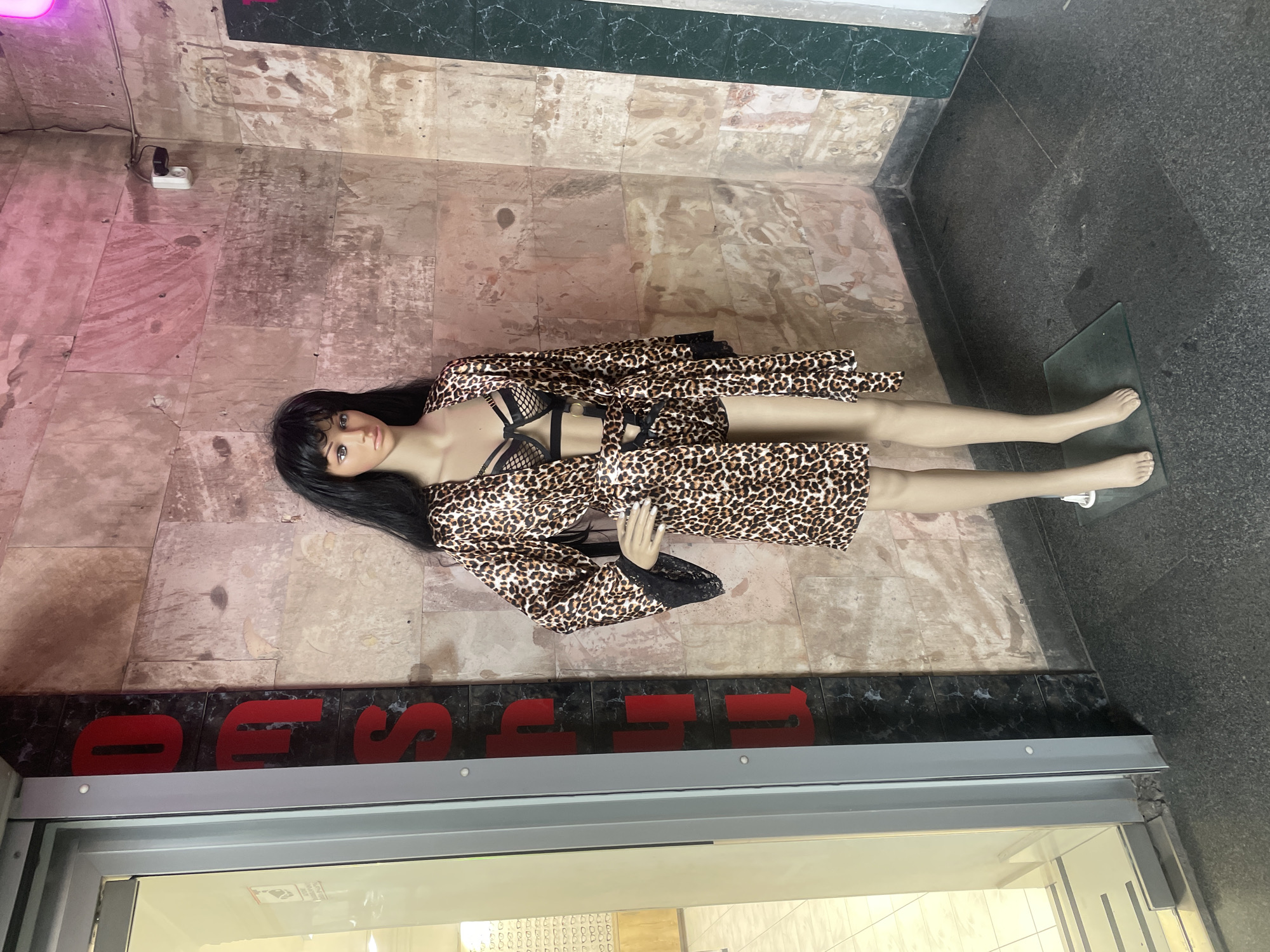 A female mannequin stands upright on a transparent glass base, positioned on a dark gray speckled floor in what appears to be a storefront entrance in Yerevan, Armenia. The mannequin has long, straight black hair, painted facial features, and is clad in a leopard print robe with black lace trim, a matching leopard print short-shorts, and a black mesh-and-strap bra with metallic rings. Its left hand rests on its hip.
To the left of the mannequin is a glass door with a gray frame, leading into an interior space with white tiled floors and a display of eyeglasses on the far wall. Adjacent to the door, a black vertical panel displays large, red, partially visible letters that spell "DOS". The background wall behind the mannequin consists of light brown and tan stone tiles exhibiting wear and pinkish lighting hues. A white security camera is mounted on this wall in the upper right quadrant of the scene. A vertical strip of dark green marble-like paneling marks the corner of the wall to the right.