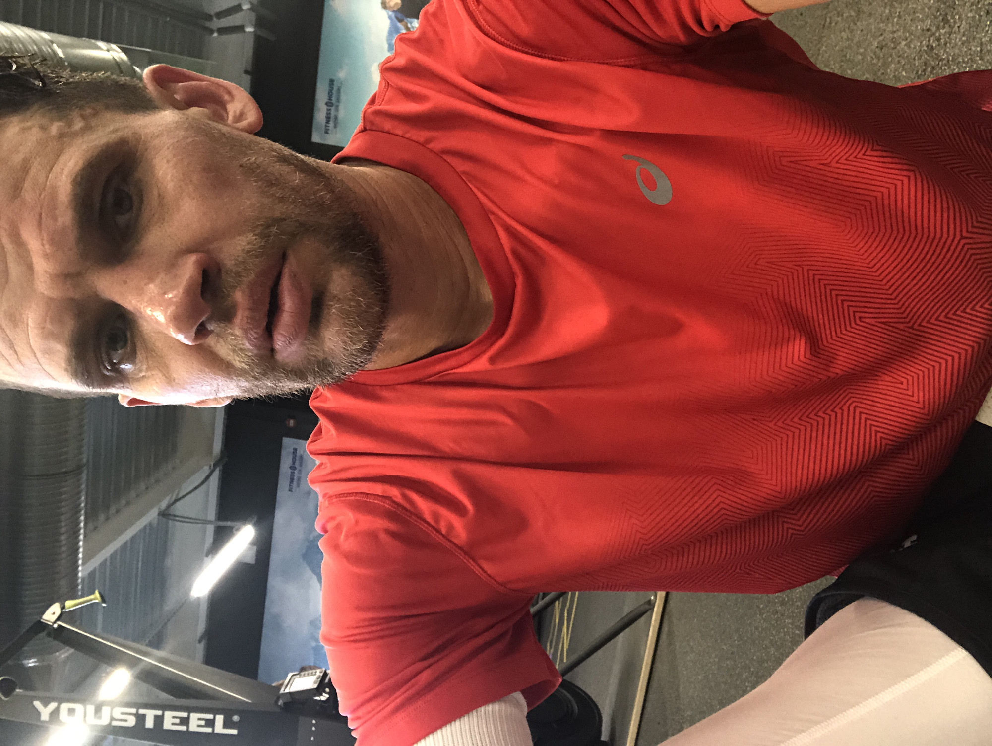 A man is taking a selfie in a gym. He is wearing a red t-shirt and white compression sleeves. He has a beard and is sweating. In the background, there is gym equipment, including a rowing machine, and a mural on the wall. There is no indication that this image is from Saint Petersburg, Russia.