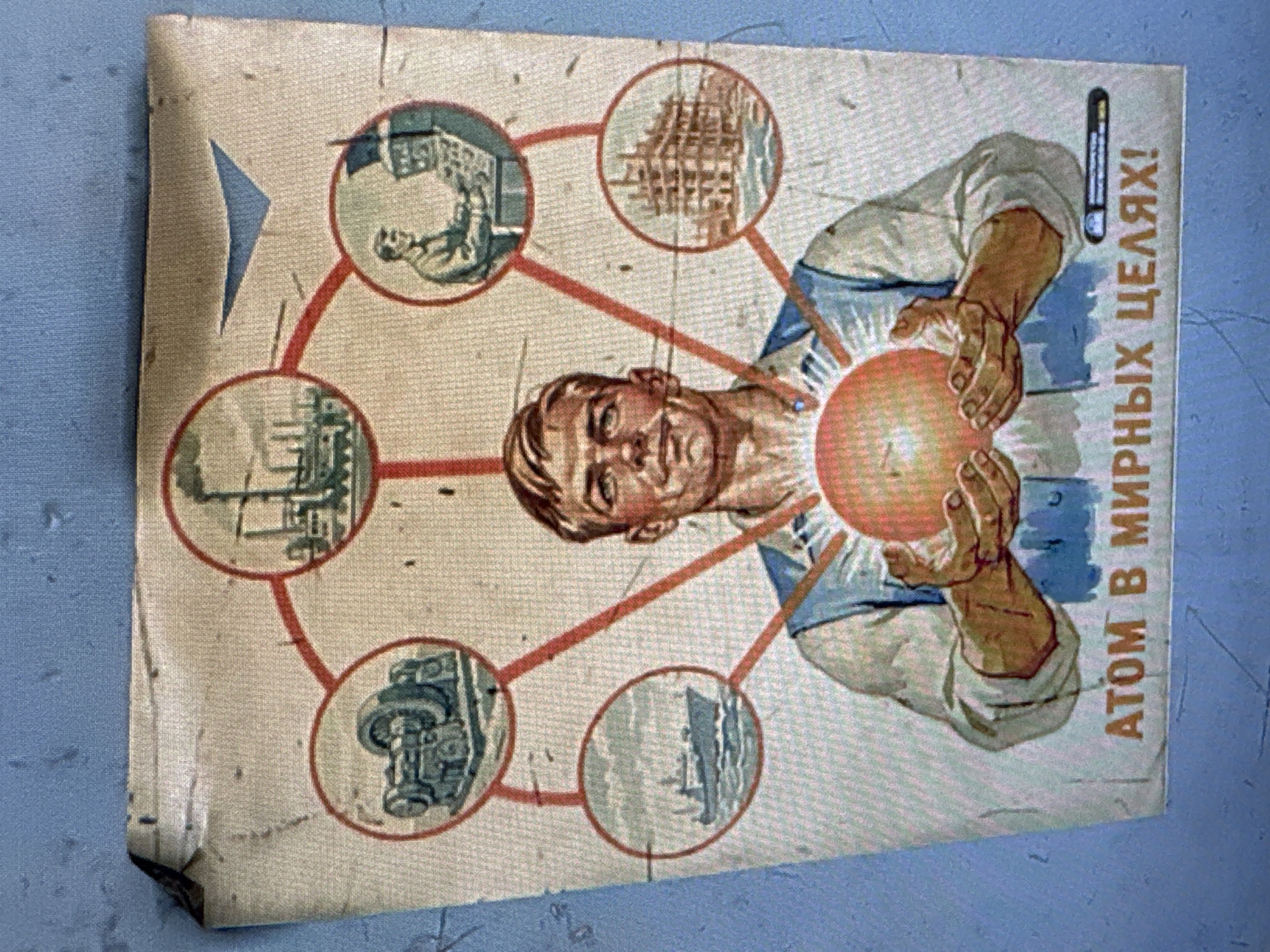 A weathered, portrait-oriented Soviet-era propaganda poster is depicted, emphasizing the peaceful applications of atomic energy. The central figure is a determined-looking man, presented from the chest up, who holds a glowing orange sphere, symbolizing an atom or nuclear power, between his hands. He is dressed in a light-colored shirt with blue accents, possibly a worker's or scientist's attire.
Radiating outwards from the glowing sphere are six red lines, each connecting to a circular vignette that illustrates a different beneficial use of atomic energy. These vignettes include:
1. An industrial worker or technician operating controls inside a facility.
2. A construction site featuring scaffolding and building structures.
3. A factory or power plant with multiple smokestacks, signifying industrial production.
4. Ships or boats navigating water, representing maritime transportation or industry.
5. Agricultural machinery, specifically a tractor plowing a field.
6. Another industrial scene or urban development with prominent buildings.
The poster's background is a light, textured off-white, and it shows signs of age, including creases, minor tears (notably at the bottom left corner), and general wear. Along the right side of the poster, oriented vertically, is the Russian text: "АТОМ В МИРНЫХ ЦЕЛЯХ!", which translates to "ATOM FOR PEACEFUL PURPOSES!". The overall tone is one of optimism and progress through technology. The poster is photographed against a blue-grey, textured surface, likely indoors in Katwijk, Netherlands.