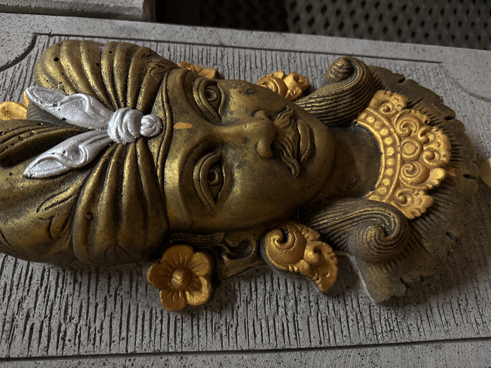 A close-up, medium shot, eye-level, outdoor, low-angle, vertical, detailed, realistic, and bright image features a gold-colored, carved stone relief of a man's face. The relief is attached to a textured, grey stone wall. The man wears a gold turban with a silver embellishment on the front. He has a mustache and a detailed gold necklace. A gold flower is visible on the left side of his head, and his hair is styled in curls. The background is slightly blurred, showing a dark, patterned surface on the right and the textured wall on the left. The image appears to be taken at a temple or cultural site in Kuta, Indonesia.
