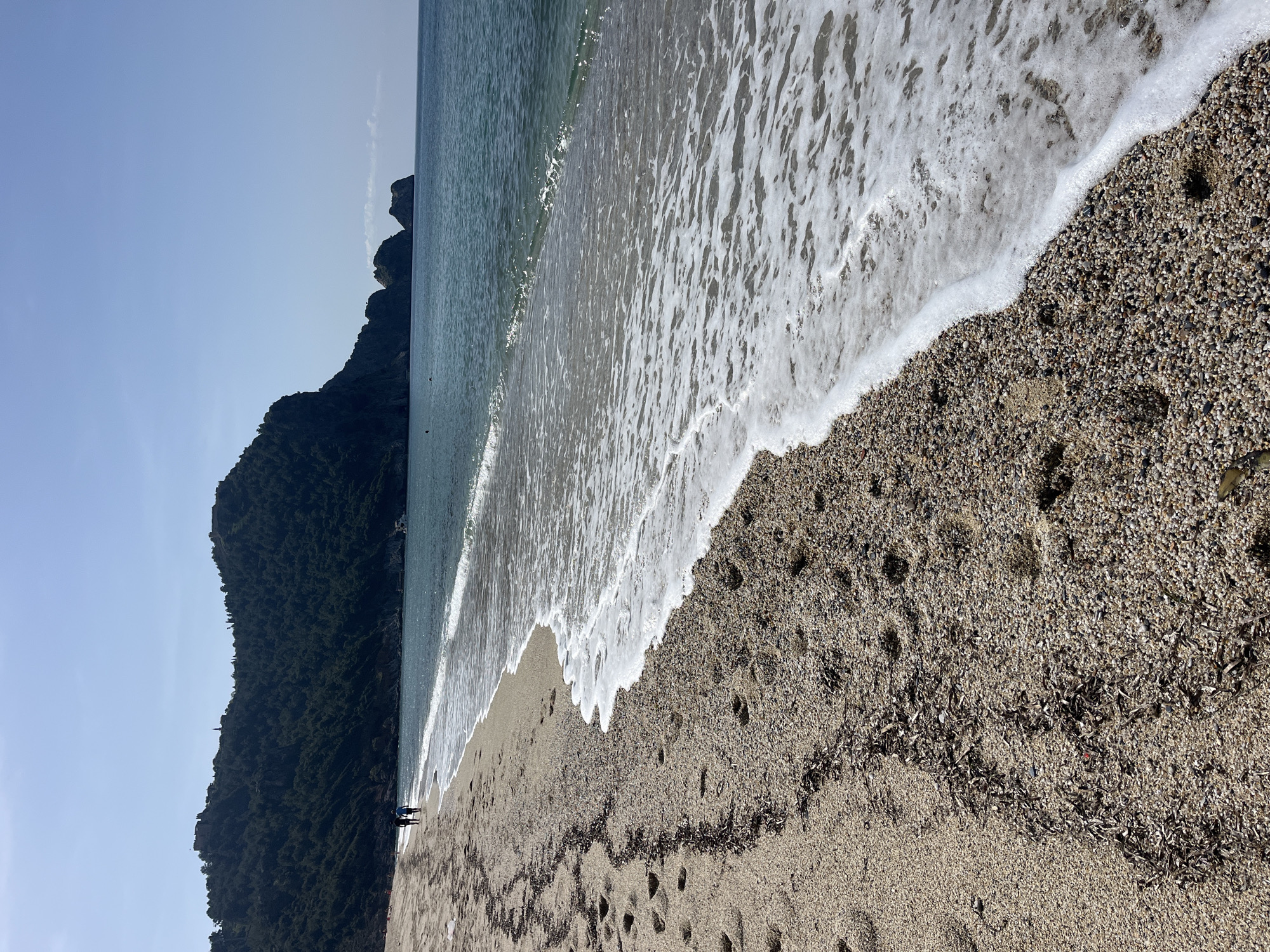 The scene is a beach in Alanya, Turkey. The image shows a rocky cliffside covered in vegetation on the left side of the frame, bordering the coastline. The beach itself is composed of coarse sand and small pebbles. The water's edge features foamy waves washing onto the shore. There are a few dark clumps of seaweed scattered on the sand. Two people can be seen standing near the cliffside, near the vegetation. The sky is light blue.