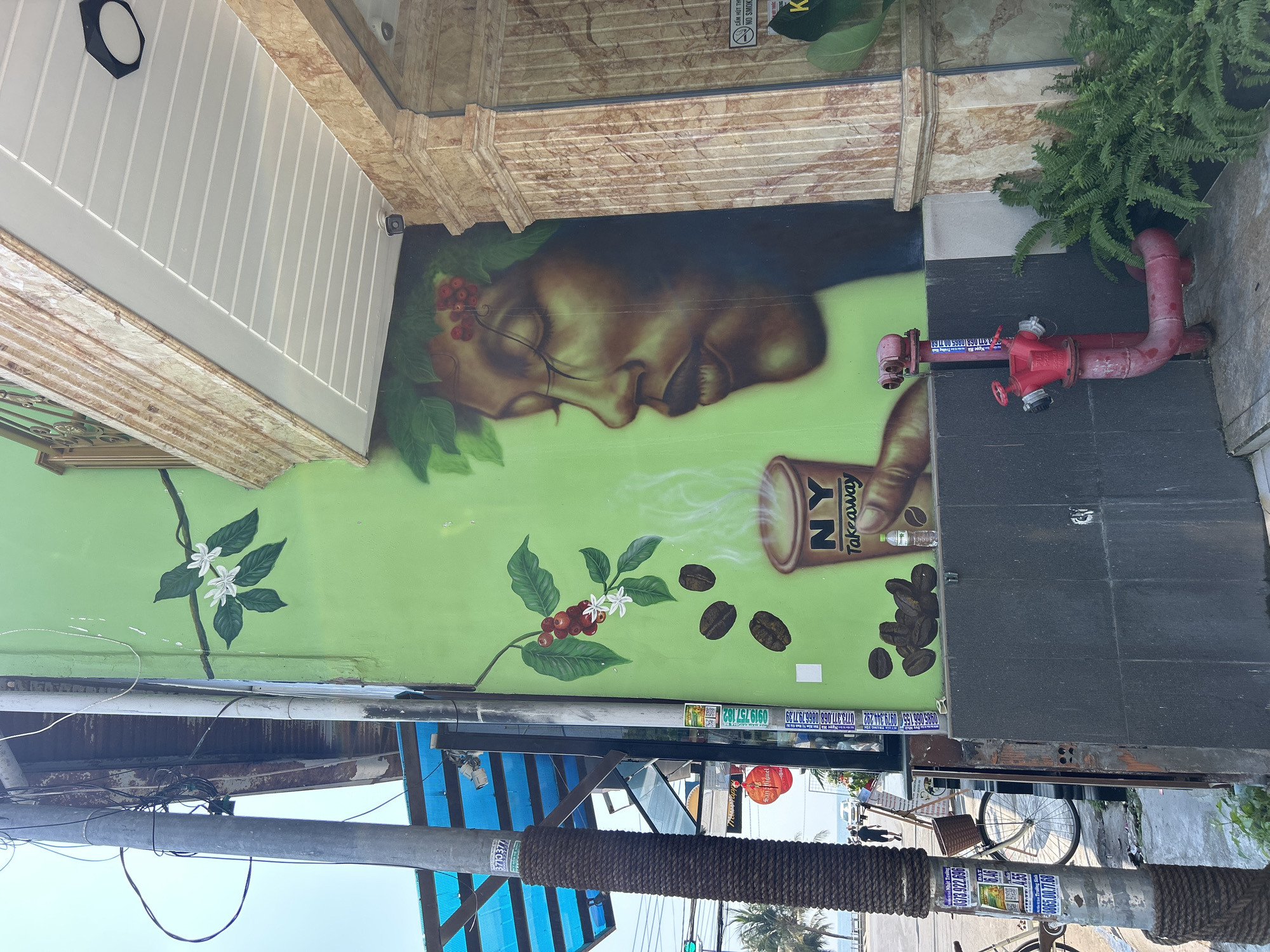 The image shows a mural in Bắc Nha Trang, Vietnam. The mural, painted on a light green wall, features the face of a person with closed eyes adorned with coffee berries, with a cup of "NY Takeaway" coffee next to them. Coffee beans are scattered around the cup. Above the mural, there's a building facade with marble accents and white panels. Next to the mural, a red fire hydrant sits on a dark grey tiled surface. The foreground shows a utility pole wrapped in a brown rope, with various advertisements and phone numbers displayed. A bicycle is parked nearby. Overhead, there are utility wires and glimpses of the sky.
