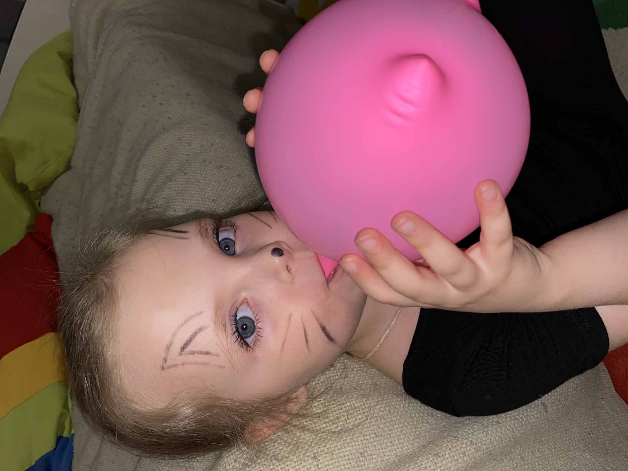 A young child with light hair and blue eyes lies on a textured beige surface, possibly a blanket or cushion. The child has dark markings on their face, resembling cat whiskers and an arrow shape on the forehead. They are positioned in a reclined position, angled towards the camera. The child is blowing up a pink balloon, which is held in their hands. The child wears a black top and a thin silver necklace. Other surfaces in the background include green and red-colored fabrics. There is no indication in the image of the location being Votkinsk, Russia.