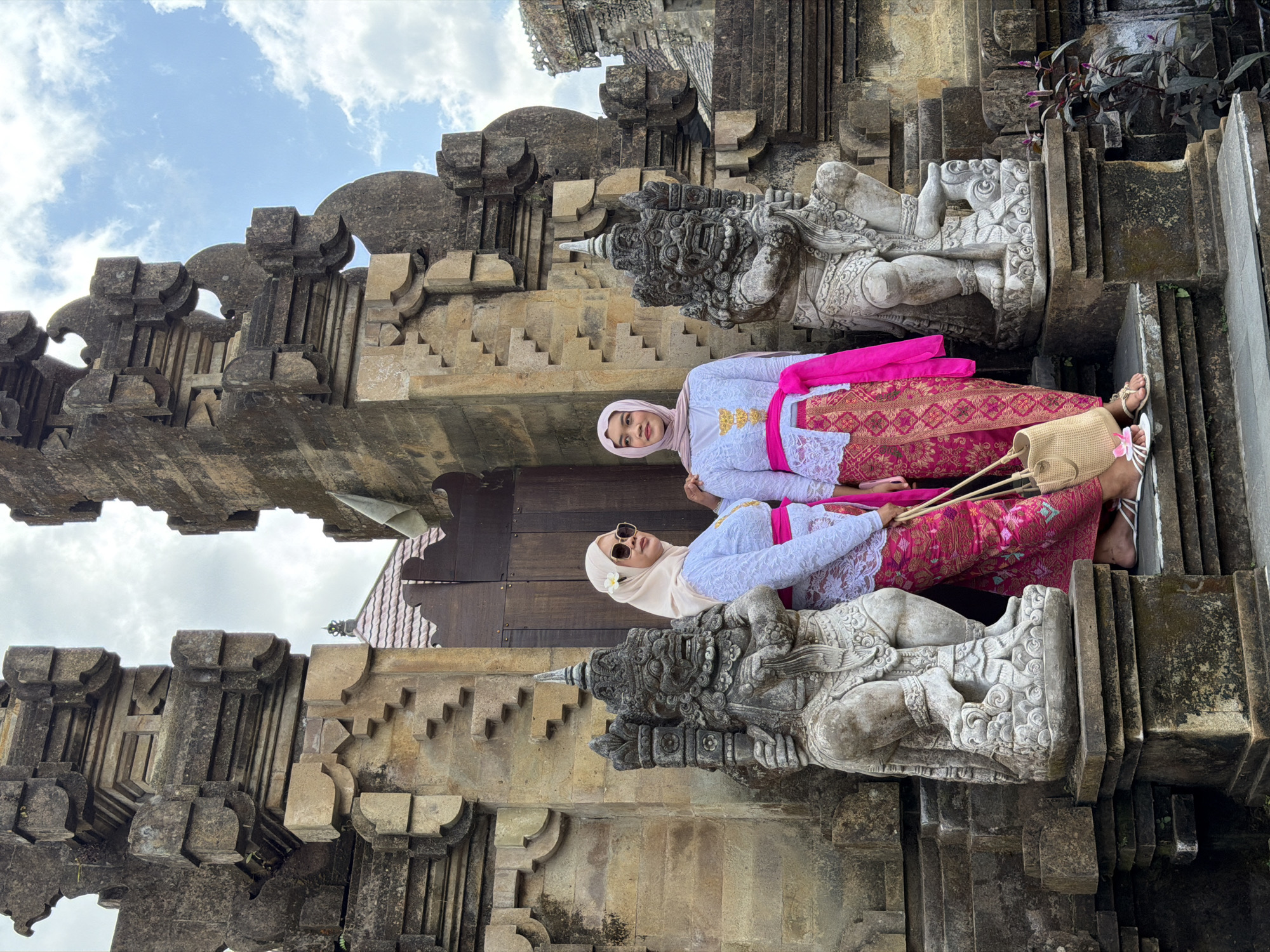 Two individuals wearing traditional Balinese attire stand in front of an ancient stone structure with intricate carvings. The structure appears to be a temple gateway, characterized by large, weathered stone pillars adorned with mythical figures. The individuals are dressed in white lace tops and colorful sarongs with a bright pink sash tied around their waists. One individual wears sunglasses and a beige headscarf with a flower tucked behind her ear, while the other wears a pink headscarf. Both are holding woven handbags. The background shows a cloudy sky with hints of blue and a glimpse of a red-tiled roof. The location is identified as the Pura Penataran Agung Susut temple in Susut, Bali, Indonesia.
