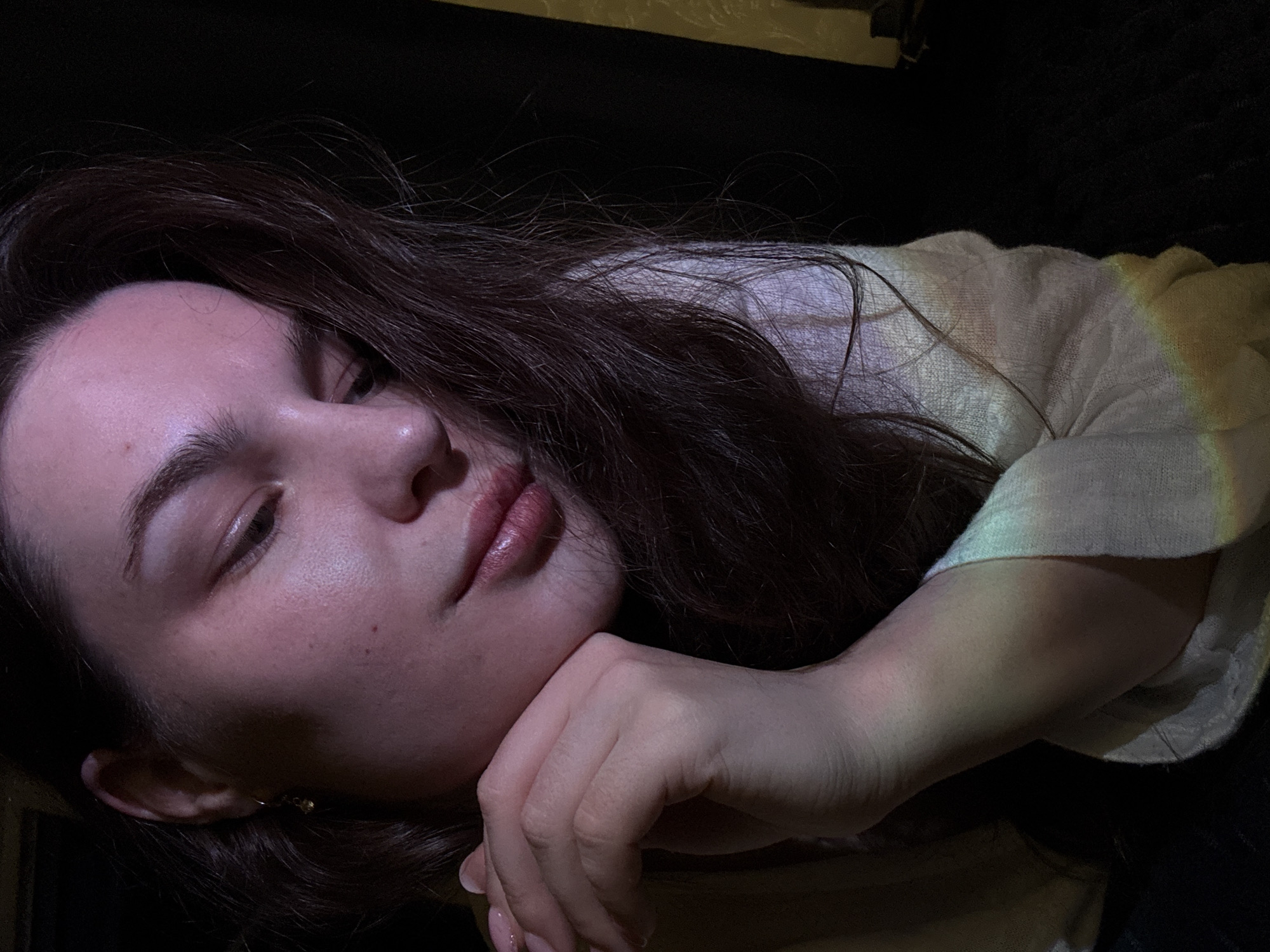 The image is a close-up, dimly lit, indoor shot of a young woman's face and upper body. She has dark hair, fair skin, and is looking down and to her right, with her chin resting on her hand. She is wearing a light-colored, possibly linen, top. There is a faint rainbow-like light cast on her clothing and skin, likely from a light source. The background is dark and indistinct. The location of this image in Chaikovsky, Russia, is not discernible from the visual information provided.