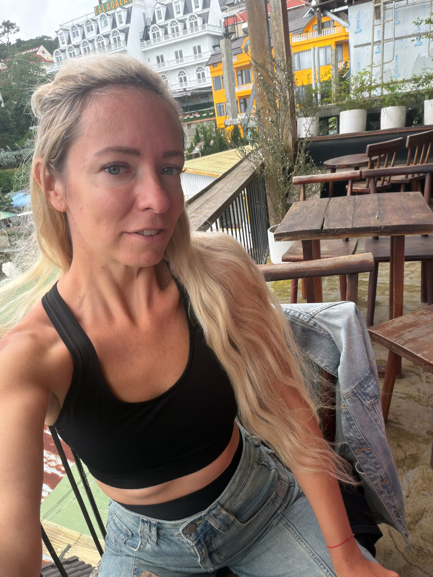 A blonde woman with long, wavy hair is taking a selfie. She is wearing a black sports bra, denim shorts, and a denim jacket draped over her shoulder. Her hair is pulled back in a messy bun, with some strands falling loose around her face. Her expression is relaxed, with a slight smile.
The woman is seated at an outdoor cafe with rustic wooden tables and chairs. In the background, there is a multi-story building with white architecture and dark roofs, possibly a hotel or resort, with the word "DALADALAT" visible on its facade. Next to it, a bright yellow building is also visible. The cafe appears to be located on a hillside overlooking the city of Da Lat, Vietnam, as indicated by the architecture and the implied elevation. The environment suggests a scenic overlook with a view of the city.