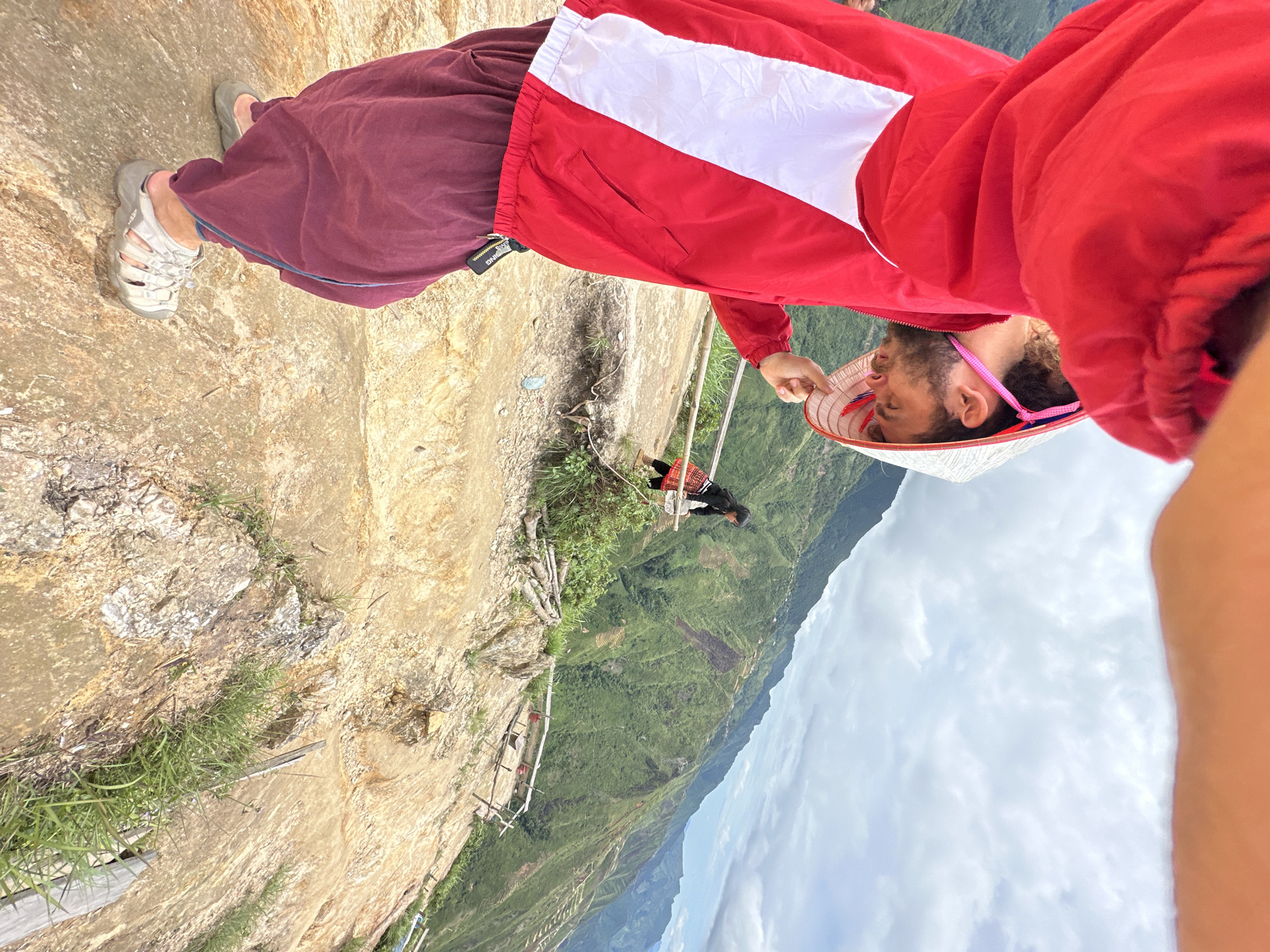 In the city of Ta Xua, Vietnam, a person wearing a red jacket, maroon pants, and sandals is holding a conical hat. The person is on a rocky, mountainous terrain, with a second person visible in the background wearing a black shirt and a red and white patterned skirt. The environment consists of lush green mountains with terraced fields under a cloudy sky. A wooden railing is present in the foreground.