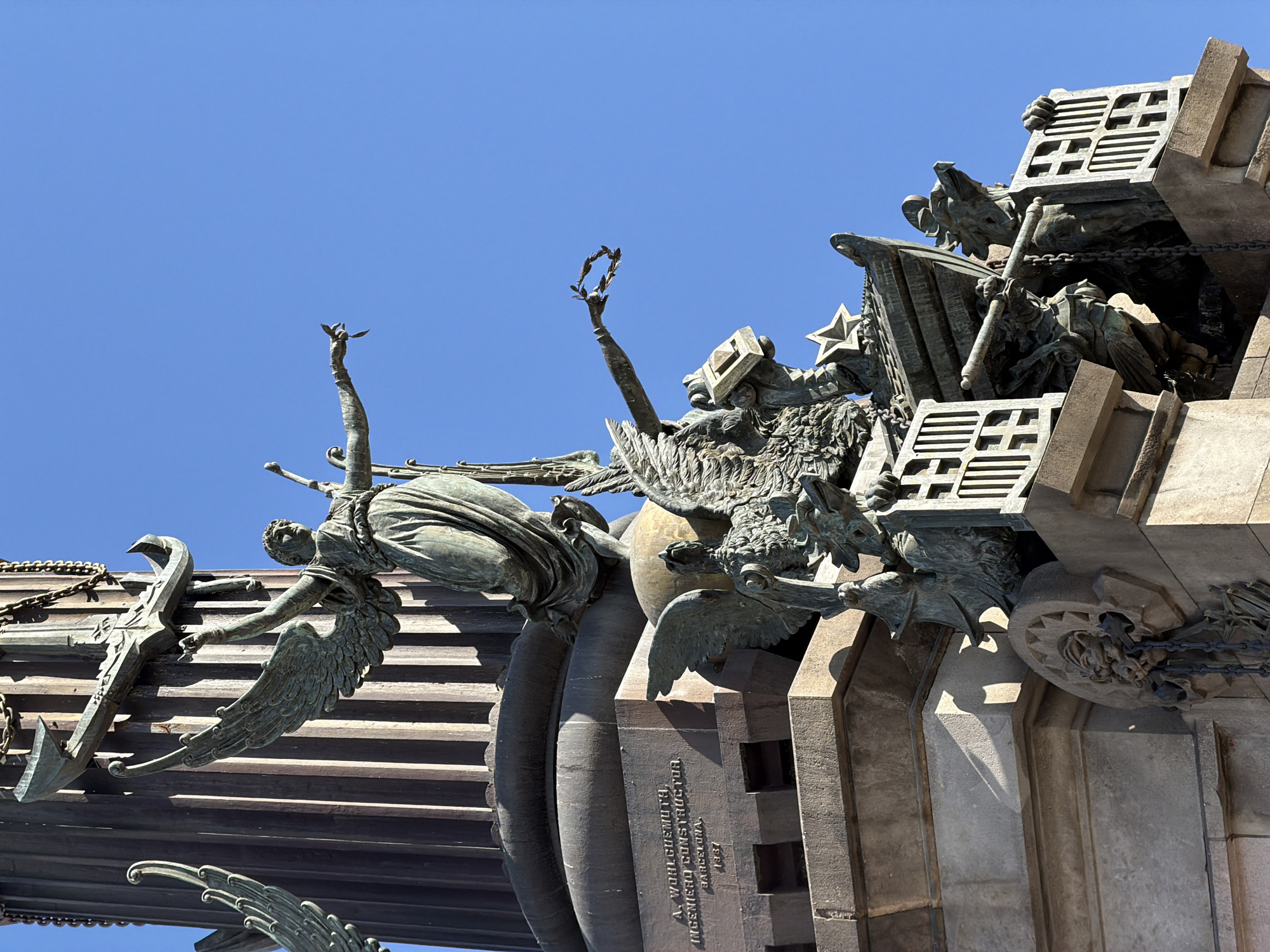 This is a close-up, low-angle shot of a monument against a clear blue sky. The monument is adorned with bronze sculptures of winged figures and architectural elements. In the foreground, a winged figure appears to be in mid-flight, with its arms outstretched, grasping what looks like an anchor or a large, ornate key. Another winged figure is visible in the upper part of the image, holding a laurel wreath. Behind these figures, more bronze sculptures are present, including what appears to be a star and elements resembling an eagle's head or a dragon's face. The monument itself is made of stone and features intricate carvings and metal chains.
Visible text on a stone section of the monument reads: "A. WOHLGEMUTH, INGENIERO CONSTRUCTOR, BARCELONA, 1931". The bright sunlight and sharp shadows suggest that it is a clear, sunny day, likely midday or early afternoon. The monument's style and the inscription mentioning "Barcelona" strongly indicate that the location is Barcelona, Spain. The overall impression is one of grandeur and historical significance, with the detailed sculptures conveying a sense of power and aspiration.