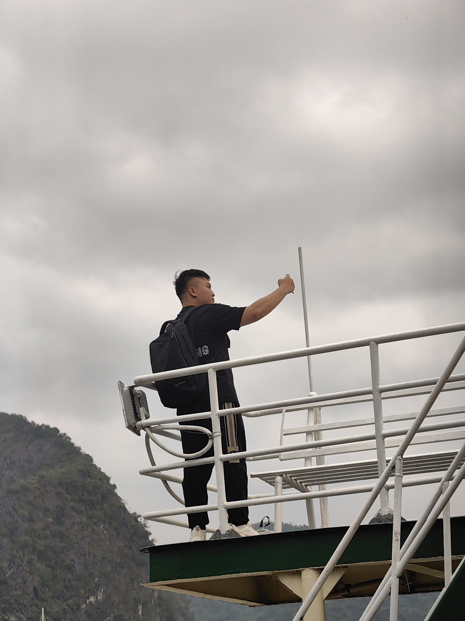 A person stands on a platform surrounded by white railings, possibly on a boat or structure overlooking water, and holds up a small white object in their right hand, against a background of cloudy skies. The person wears black clothing and a black backpack. A hillside with greenery is visible in the background. The location is in the city of Hải Phòng, Vietnam.