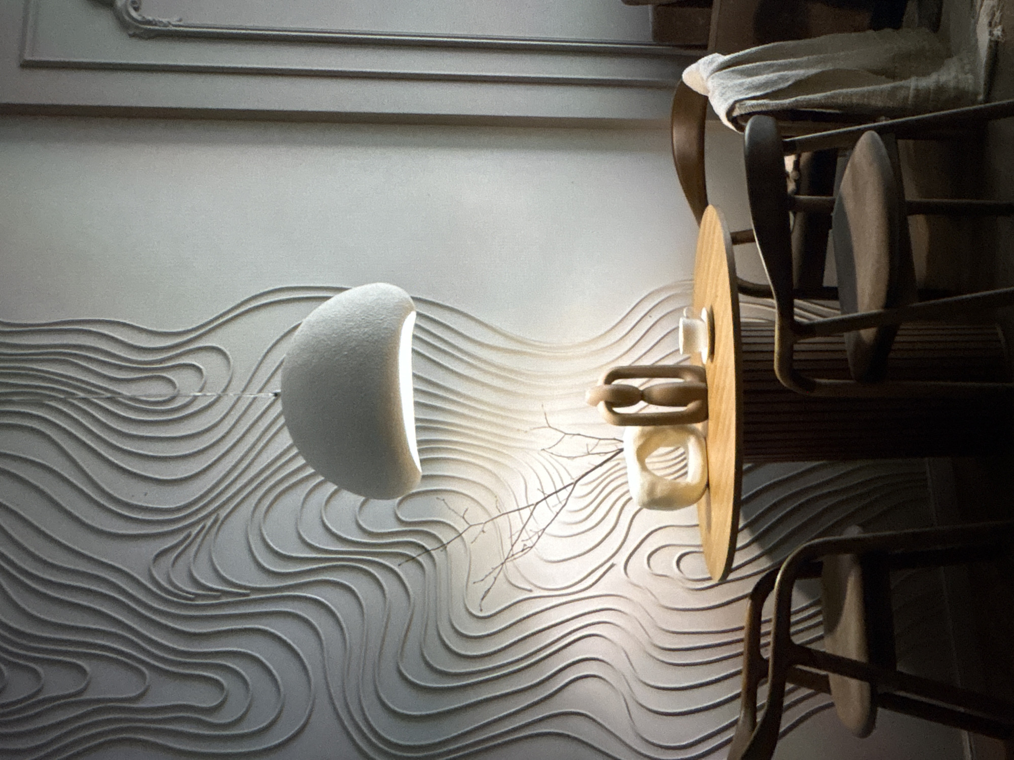 A bright room features a textured wall with horizontal wave patterns, upon which a half-sphere lamp is mounted. A wooden table with a cylindrical base and two wooden chairs are positioned to the right, with a white cloth draped over one. A decorative branch sits on the table. The location is stated to be Ембаево, Russia.